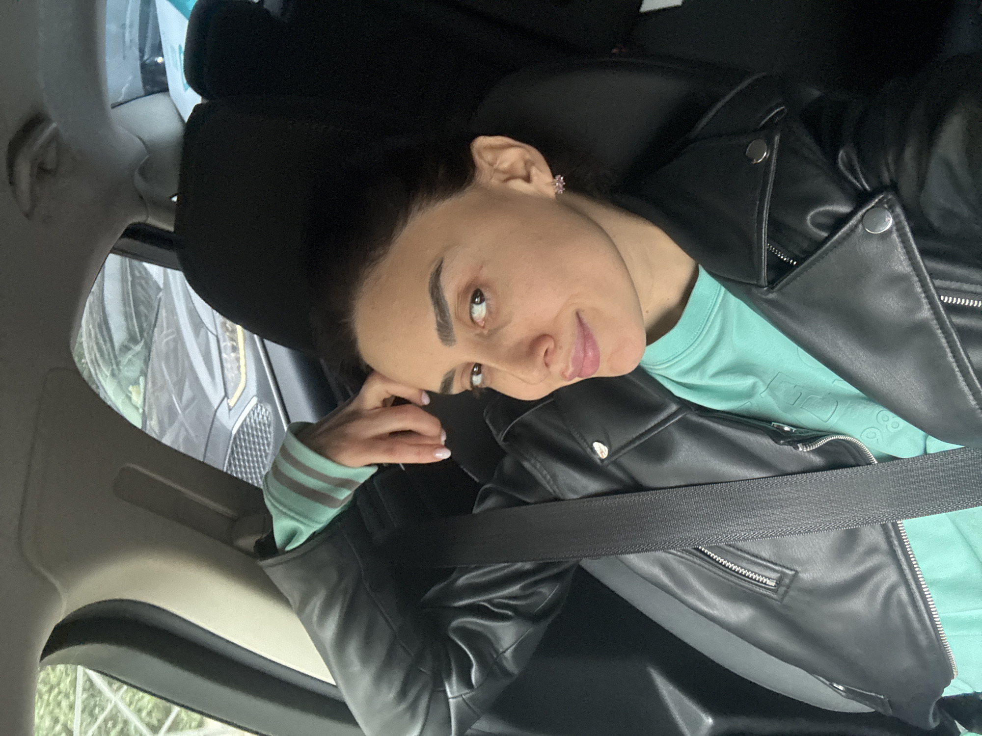 A portrait-oriented photo captures a young woman seated in what appears to be the front passenger seat of a car, likely in Tbilisi, Georgia. She is looking upwards and to her left (viewer's right) with a slight, relaxed smile. Her left hand is casually resting near her temple.
The woman has dark hair pulled back, prominent dark eyebrows, and brown eyes. She is wearing a black leather moto jacket over a mint green long-sleeved top. The jacket features silver snaps on the lapels and visible zippers. Her mint green top has horizontal stripes on the cuffs, and a faint, indistinguishable raised pattern or text is visible on its front. A black seatbelt is securely fastened across her chest and lap. In her right ear, she wears a small, pink, possibly floral or star-shaped earring. Her fingernails have a light, neutral polish.
The car's interior is visible, including a dark headrest, parts of the ceiling, and a window frame. Outside the car window, elements of another dark-colored vehicle and some blurred greenery, likely trees or bushes, can be seen, indicating a daytime setting. The overall mood of the image is calm and casual. No clear, legible text is visible in the image.