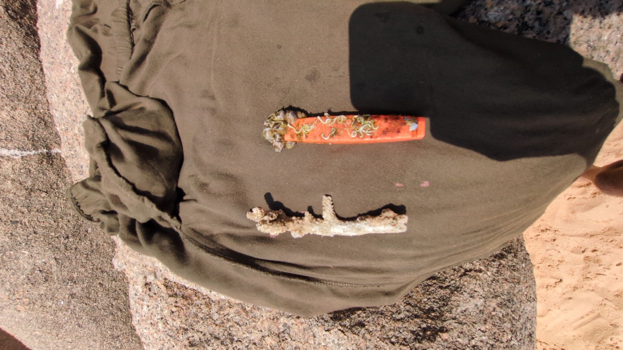 A close-up, high-angle shot shows an olive-green t-shirt laid on a rough, textured granite surface. Superimposed on the shirt are two objects: An orange, oblong item, possibly a lighter or small tool, with marine encrustations and small, worm-like organisms on its surface, and a piece of weathered coral. The location is likely near a rocky coastal area, possibly in or near Phuoc Dinh, Vietnam. A partial view of a person's lower leg is visible in the lower right corner of the shot, resting on a sandy surface. The environment appears to be outdoors, with sunlight casting shadows on the scene.