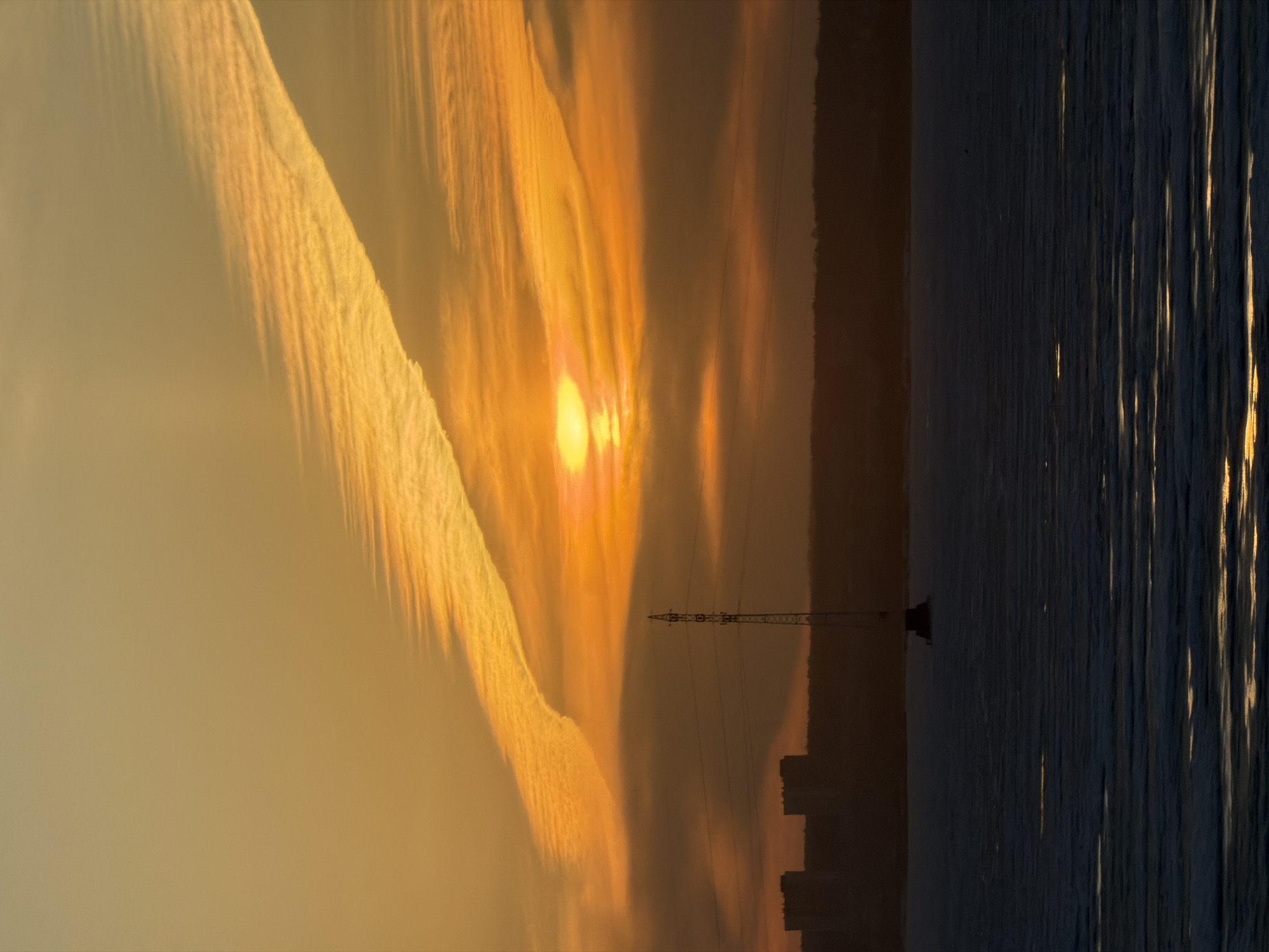 The image depicts a sunset over a body of water, with buildings and a transmission tower silhouetted against the sky. The sky is filled with clouds, with contrails. The water reflects the light, creating bright streaks. The location is Izhevsk, Russia.