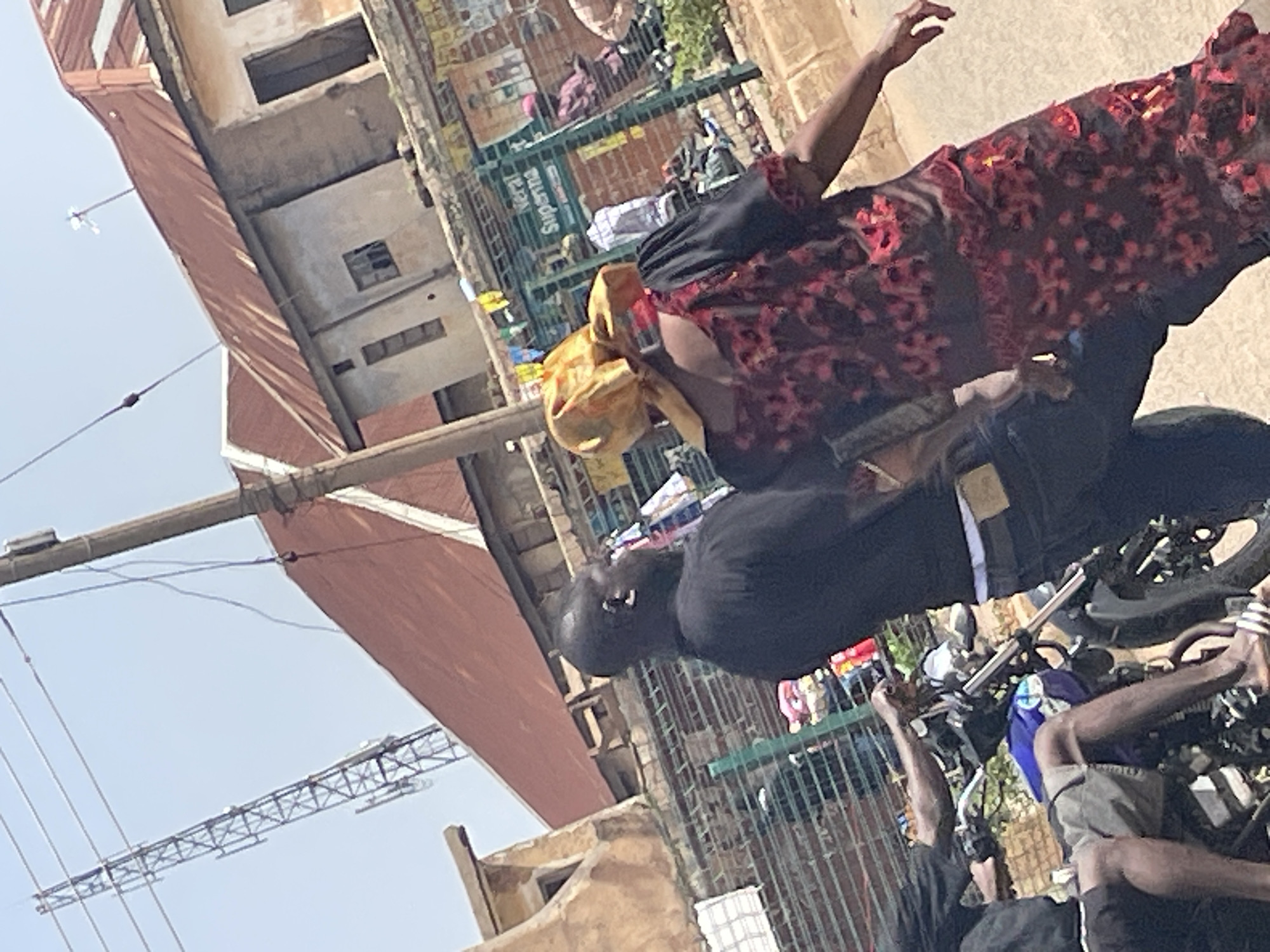 In Bukuru, Nigeria, on a sun-drenched day, two individuals walk on a dusty street. A man, seen from the back, wears a black shirt and dark trousers, his left ear visible. Beside him, a woman in a red and black patterned dress and a yellow headscarf walks ahead, her left arm extended. In the foreground, a motorcycle is partially visible, with a person's legs and feet wearing sandals on its left side. The background reveals urban architecture, including buildings with rust-colored tin roofs, a tall communication tower, and a market stall with various items displayed behind a green fence. The overall scene depicts a typical street environment in a Nigerian city.