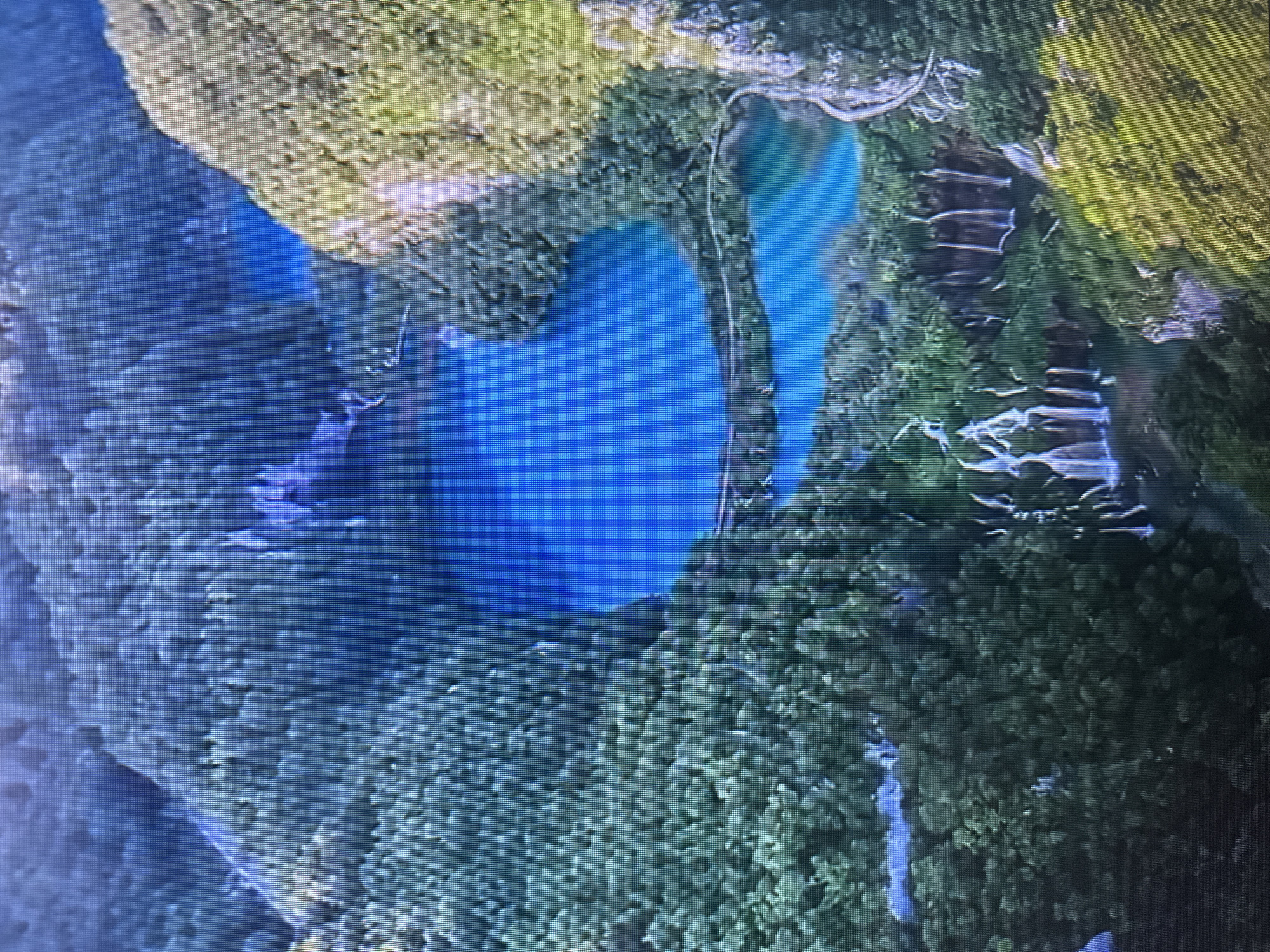 The image is an aerial view of a lush green landscape with several turquoise lakes and numerous waterfalls. The terrain is characterized by steep, forested hillsides. Wooden walkways are visible winding along the edges of the lakes and over the water. The scene depicts a natural park or reserve. The image is a photograph of a screen displaying the scene, indicated by the visible pixelation. There is no indication that this scene is located in the city of Kyiv, Ukraine; it appears to be a natural landscape.