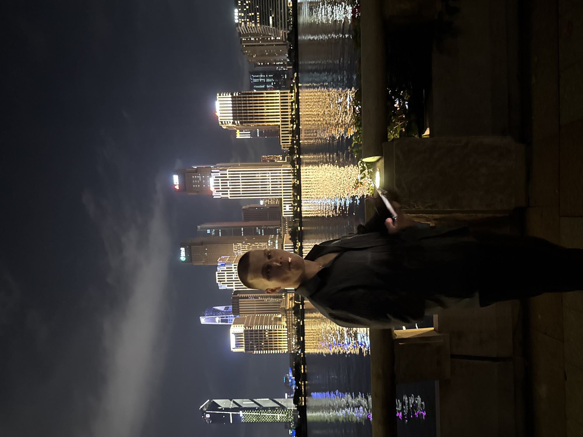 The image presents a nighttime urban scene in Haizhu District, China, with a male individual positioned in the foreground. The man, with short dark hair and fair skin, wears a dark collared shirt, revealing a thin gold-colored chain around his neck. He holds a white cylindrical object, consistent with a vape or cigarette, to his mouth with his right hand, while his left hand holds an out-of-focus smartphone. He stands in front of a light-colored stone railing, topped with white flowers and foliage illuminated by a small light. Beyond the railing lies a dark body of water, reflecting the vibrant lights of numerous skyscrapers and commercial buildings that form an illuminated cityscape on the far bank. The city skyline features predominantly golden and white building lights, with some structures displaying blue and green accents, notably a tall building on the far left with prominent green vertical lighting. The sky above is dark with visible cloud formations, faintly illuminated by the city's ambient light.