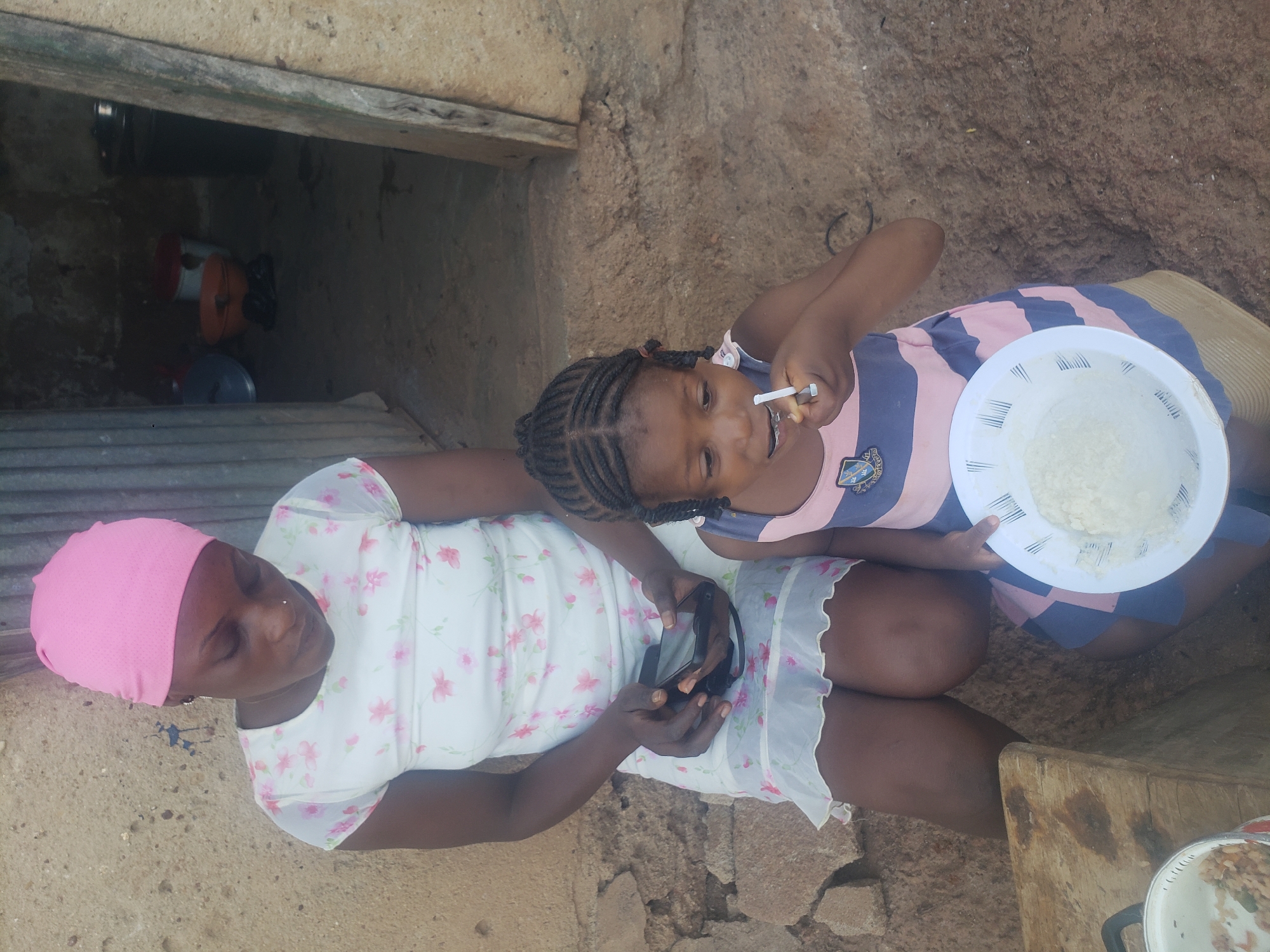 The image shows a woman and a young girl sitting outside a building in Bukuru, Nigeria. The woman is wearing a pink headscarf and a white dress with pink flowers. She is holding a black smartphone. The young girl, who is sitting to the right of the woman, is wearing a striped pink and blue dress. She is holding a spoon to her mouth and a white bowl containing food. A cooking pot is visible in the foreground to the right. The background includes a building with a dark opening and a weathered exterior. The ground appears to be uneven.
