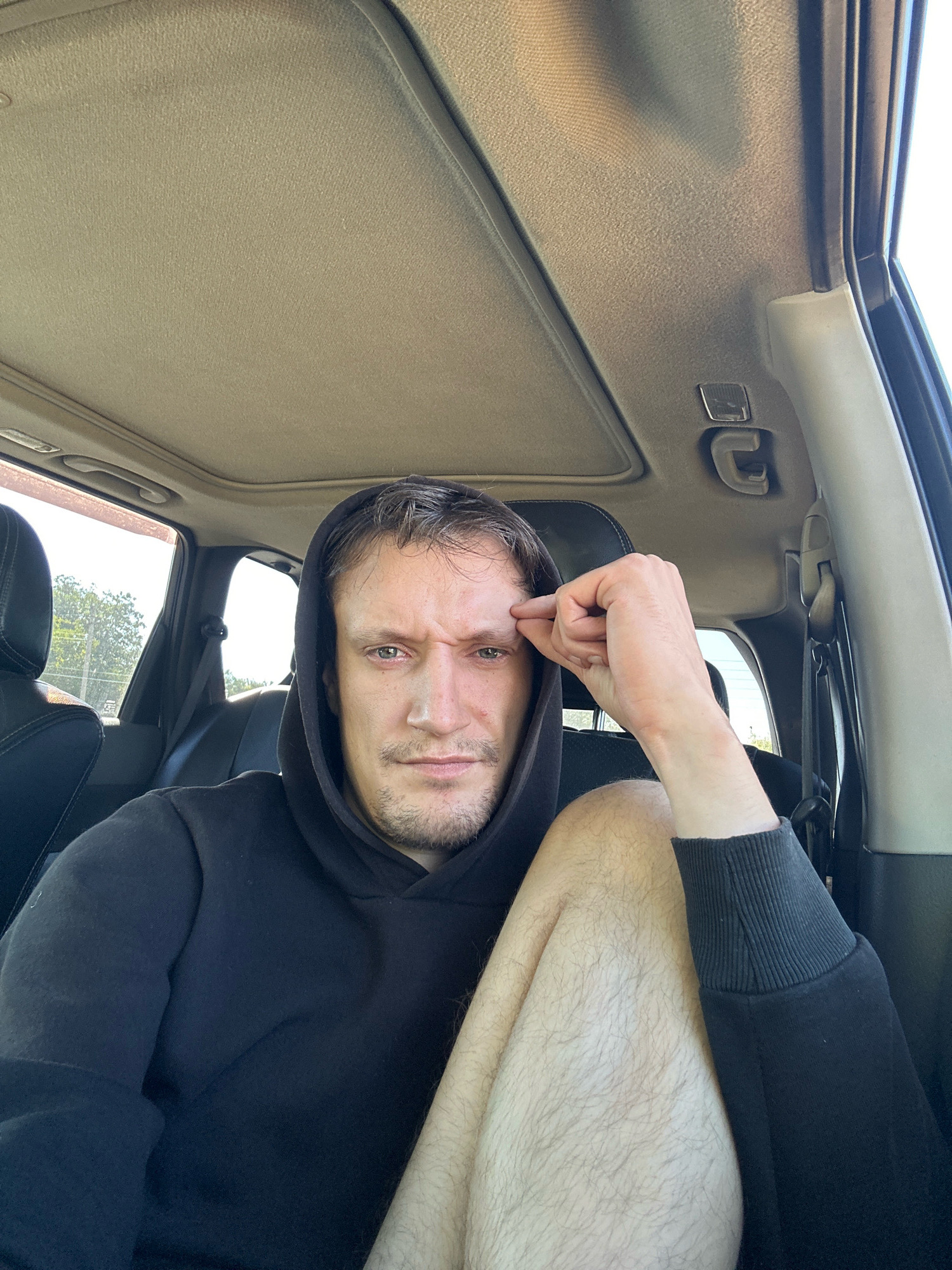 A man is sitting in the driver's seat of a car, looking at the camera. He is wearing a black hoodie and has his right hand raised to his forehead, touching his temple. The car's sunroof is open. Outside the car, trees and a pole are visible. The interior of the car appears to be black leather. The man has short, light brown hair and stubble. The image appears to be taken in Makiv, Ukraine.