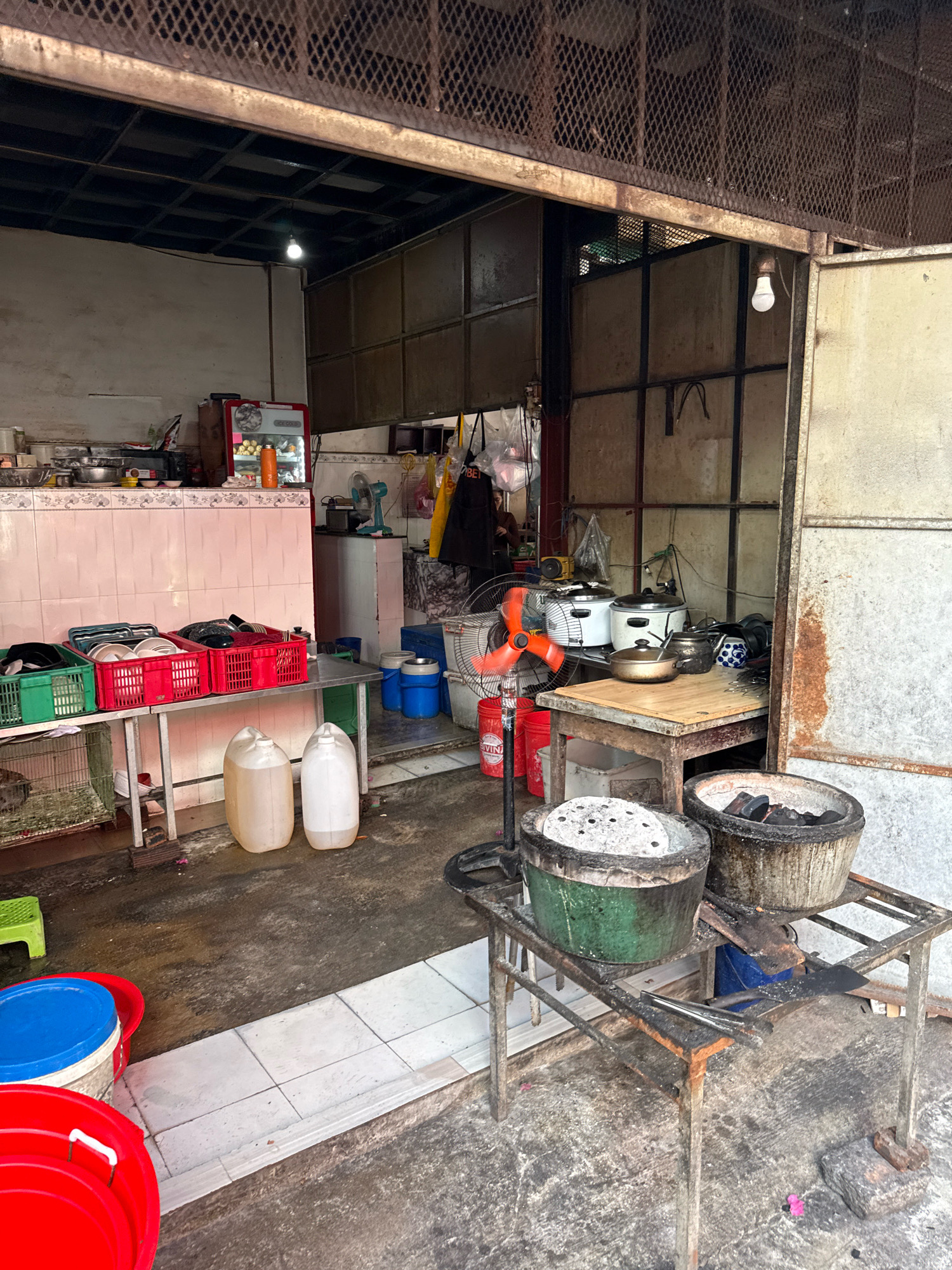 The image depicts an outdoor food stall area, likely in Nha Trang, Vietnam, characterized by a rustic and functional setup. The scene is a mix of cooking equipment and storage, suggesting an active preparation environment.
In the foreground, two large, weathered charcoal grills are positioned on a metal stand. The grill on the left contains a circular, perforated plate, while the one on the right holds burning charcoal. Various cooking utensils, including tongs, are visible on the stand. Adjacent to this, several large plastic containers, one red and one blue, are placed on the ground, indicating water or ingredient storage.
To the left, a metal counter displays an assortment of items. Green and red plastic crates hold stacks of plates and bowls. Below this counter, a wire cage contains what appears to be a live animal. Two large, translucent plastic jerrycans filled with liquid are placed on the floor near the counter.
In the mid-ground, a small red refrigerator with a glass front, displaying bottles and food items, is situated against a pink tiled wall. Various cooking appliances, including rice cookers and what looks like a kettle, are on a wooden table behind a rotating fan. Various bags and containers are also visible in this area.
The background reveals a more enclosed space with metal shelving and what appears to be a partition made of corrugated metal. The overall environment suggests a busy, functional food stall with a focus on practicality and efficiency, common in urban Vietnamese street food settings. The ground is concrete and appears wet in places, with some debris scattered around.