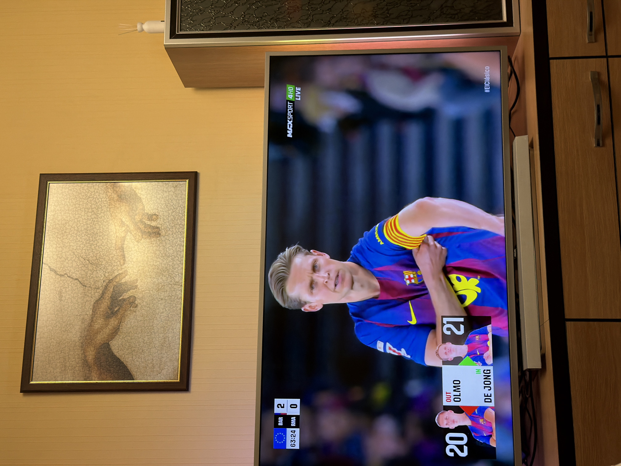 The image shows an indoor scene, dominated by a wall-mounted flat-screen television displaying a soccer (football) match. The broadcast shows FC Barcelona player Frenkie de Jong. A scoreboard indicates the score is Barcelona 2, Real Madrid 0, with a time of 63:24. The channel is Max Sport 4HD and is broadcasting live. An inset graphic indicates that Olmo has been substituted out for de Jong, wearing number 21. The hashtag "#ElClásico" is visible on the upper right of the screen.
To the left of the television hangs a framed artwork, possibly a puzzle rendition of Michelangelo's "The Creation of Adam." The wall is light yellow.
Adjacent to the TV on the right side is a set of wooden cabinets with vertical metal handles.
The image provides no information to suggest it is located in Plovdiv, Bulgaria.