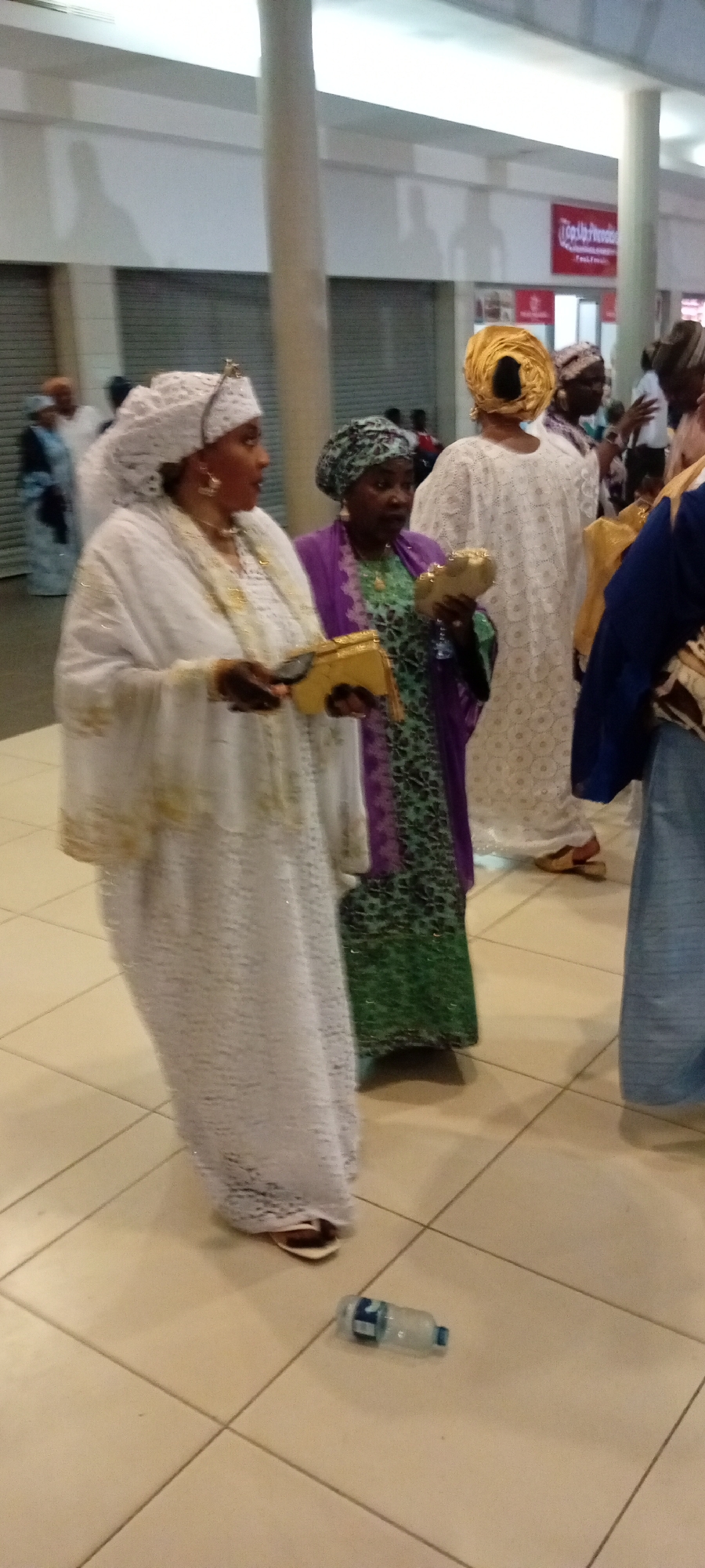 A medium shot shows three women in traditional African attire walking indoors. The woman in the foreground on the left wears a white lace dress and headscarf, holding a gold clutch. To her right, a woman in a green and black patterned dress and a purple headscarf holds a similar gold clutch. Behind them, another woman wears a white lace dress and a gold headscarf. The environment appears to be a public indoor space with tiled flooring and a sign for "Top Up Pre-Order" in the background. A plastic water bottle lies on the floor in the foreground.
