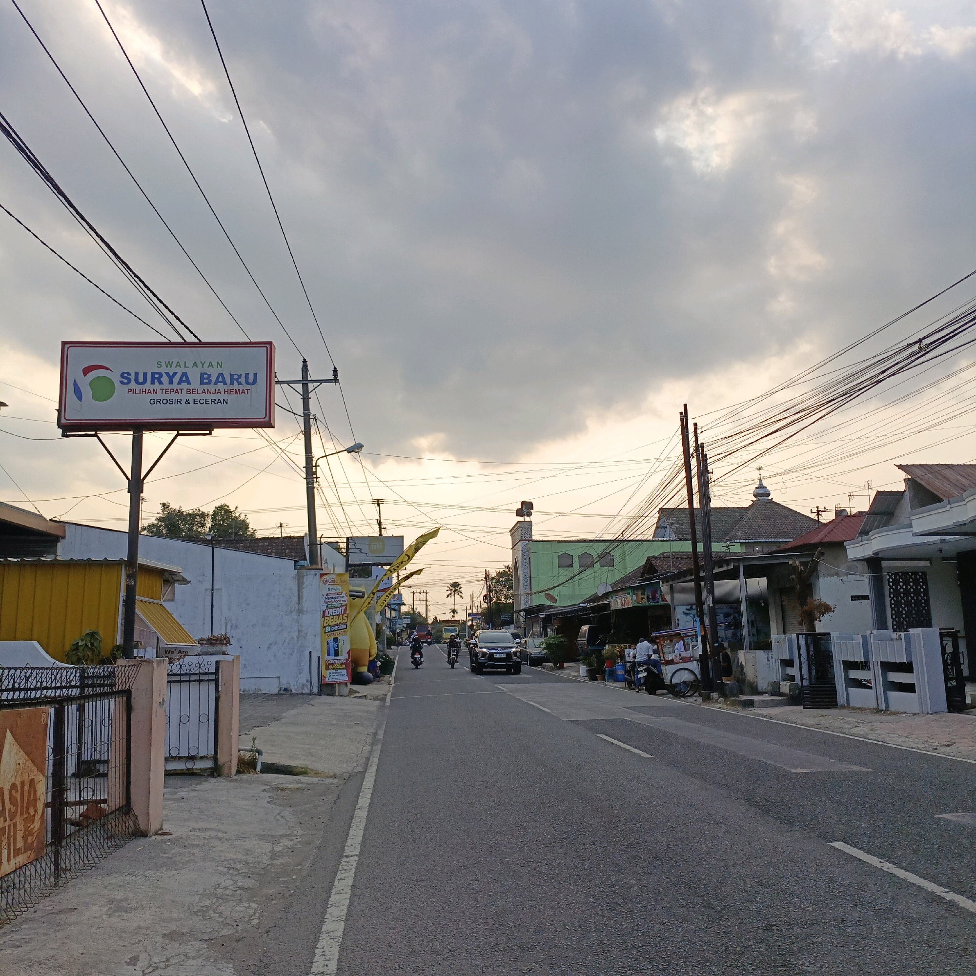 This image shows a street scene in Salatiga, Indonesia, likely during the late afternoon or early evening given the lighting and cloud cover. The foreground features a paved road with a white lane divider, and a sidewalk on the left. A prominent sign for "SWALAYAN SURYA BARU" is visible, indicating a grocery store with the slogan "PILIHAN TEPAT BELANJA HEMAT GROSIR & ECERAN". Electrical wires crisscross the sky above the street.
On the left side of the street, there's a yellow building with a fence in front of it. Further down the street, several people are seen on motorcycles and in a black SUV. Buildings line both sides of the road, including a green building with a mosque-like dome on top. Some small shops and food stalls are also present. A few people are standing or sitting near these establishments. The overall environment suggests a moderately busy urban or suburban street.