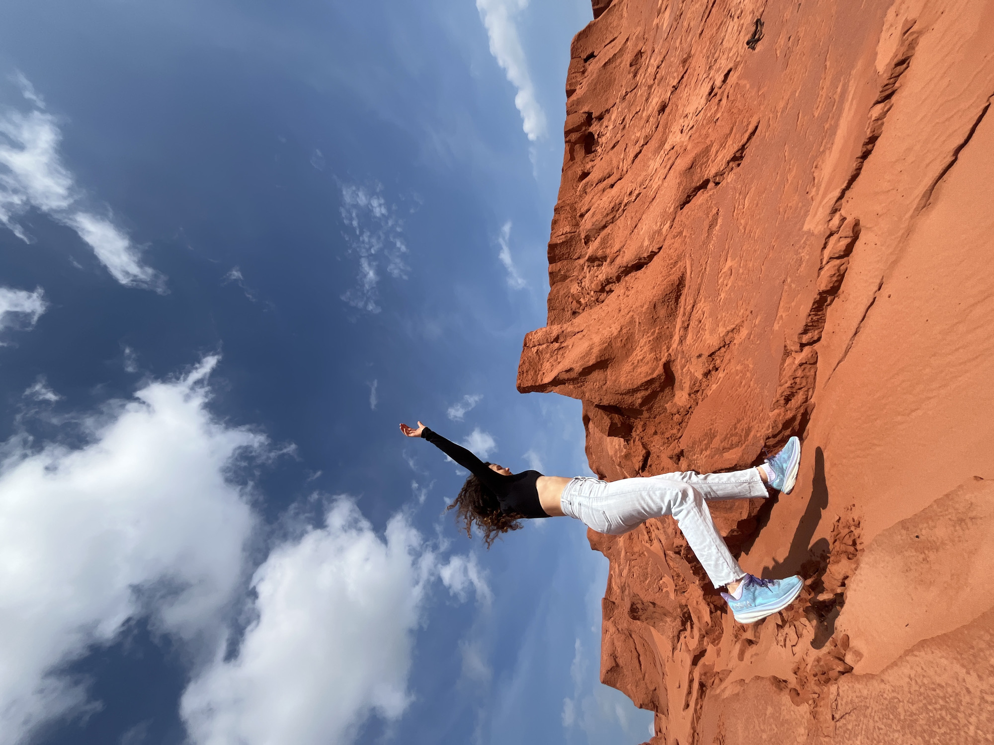 A person is captured mid-air, jumping in a desert-like landscape with red rock formations. The individual, wearing a black crop top, light-colored jeans, and blue sneakers, has their right arm raised. Their dark, curly hair is windblown, suggesting movement. The background consists of a clear blue sky with scattered white clouds, and prominent red sandstone cliffs and dunes that are characteristic of the Mui Ne sand dunes in Vietnam.