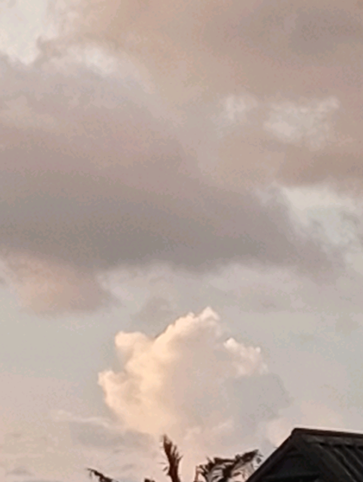 A low-angle, slightly blurred outdoor shot captures a cloudy sky with hints of pink and gray. In the foreground, the dark roof of a building juts in from the right side of the frame, partially obscuring the lower portion of the image. Above the building, silhouetted branches of trees extend upwards. A prominent, fluffy white cloud is illuminated by soft light, drawing attention in the middle ground. The overall scene appears to be from a residential area in Obosi, Nigeria, with the overcast sky suggesting a calm, possibly pre-sunset atmosphere.