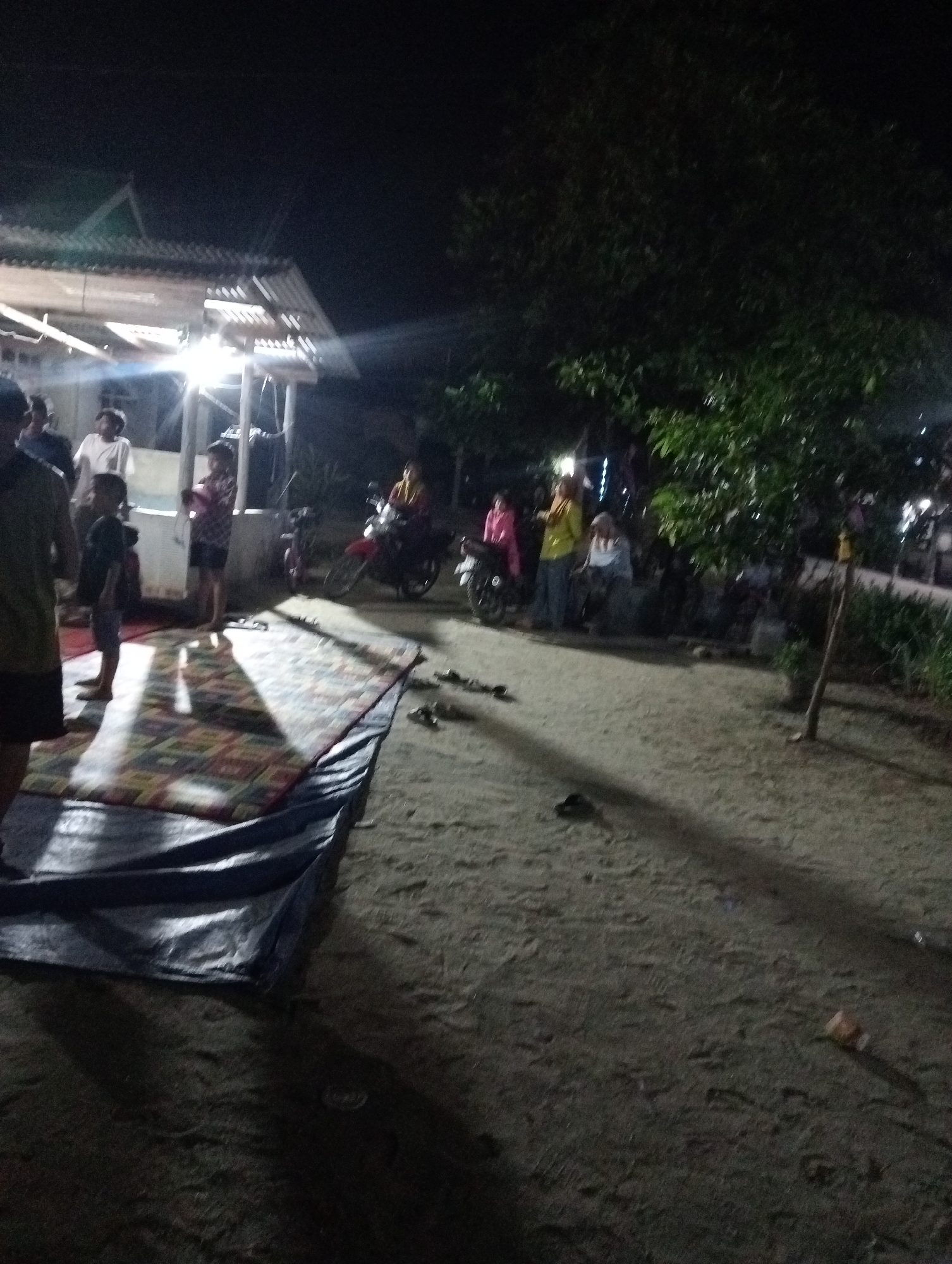 In the village of Rasuan Baru, Indonesia, at night, a group of people are gathered outdoors. A brightly lit open-sided structure, possibly a pavilion or porch, is visible in the background. Several individuals are standing near the structure, and some are on motorcycles. In the foreground, a patterned mat is laid out on the ground, partially covered by a dark tarp. The ground appears to be dirt or sand. The scene suggests a community gathering or event taking place after dark.