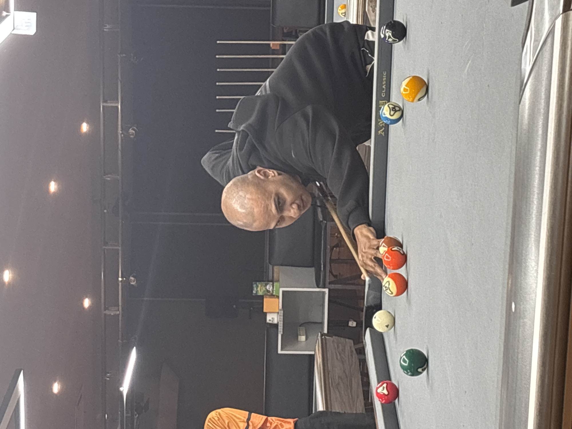A bald man wearing a black hoodie and dark pants is playing pool in a dimly lit room, possibly a bar or a billiard hall in Kuta Selatan, Indonesia. He is in the process of taking a shot, with his cue stick extended towards a group of balls on the grey felt table. Several pool balls are scattered on the table, including a red ball with the number 4, a white ball, two orange and yellow striped balls, a green ball, and a blue and yellow striped ball. Another person, wearing an orange shirt, is partially visible on the left side of the frame. The background shows a dark wall with vertical metal rods and some seating. The ceiling has multiple light fixtures.