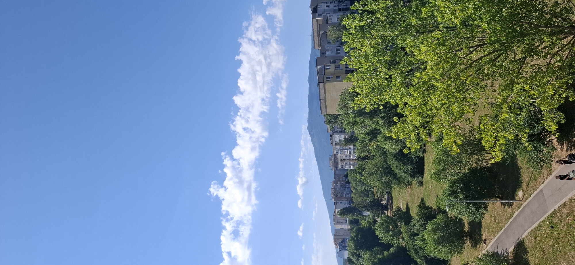The image shows a panoramic view of a city with buildings and a mountain in the background, likely taken from a high vantage point in Sofia, Bulgaria. The sky is a clear blue with scattered white clouds. In the foreground, there is a park-like area with lush green trees and a paved pathway. Two people are walking on the pathway, one pushing a stroller. Behind the park, a row of apartment buildings can be seen, and further in the distance, a mountain range looms under the sky. The overall scene depicts a sunny day in an urban environment with a significant amount of green space.