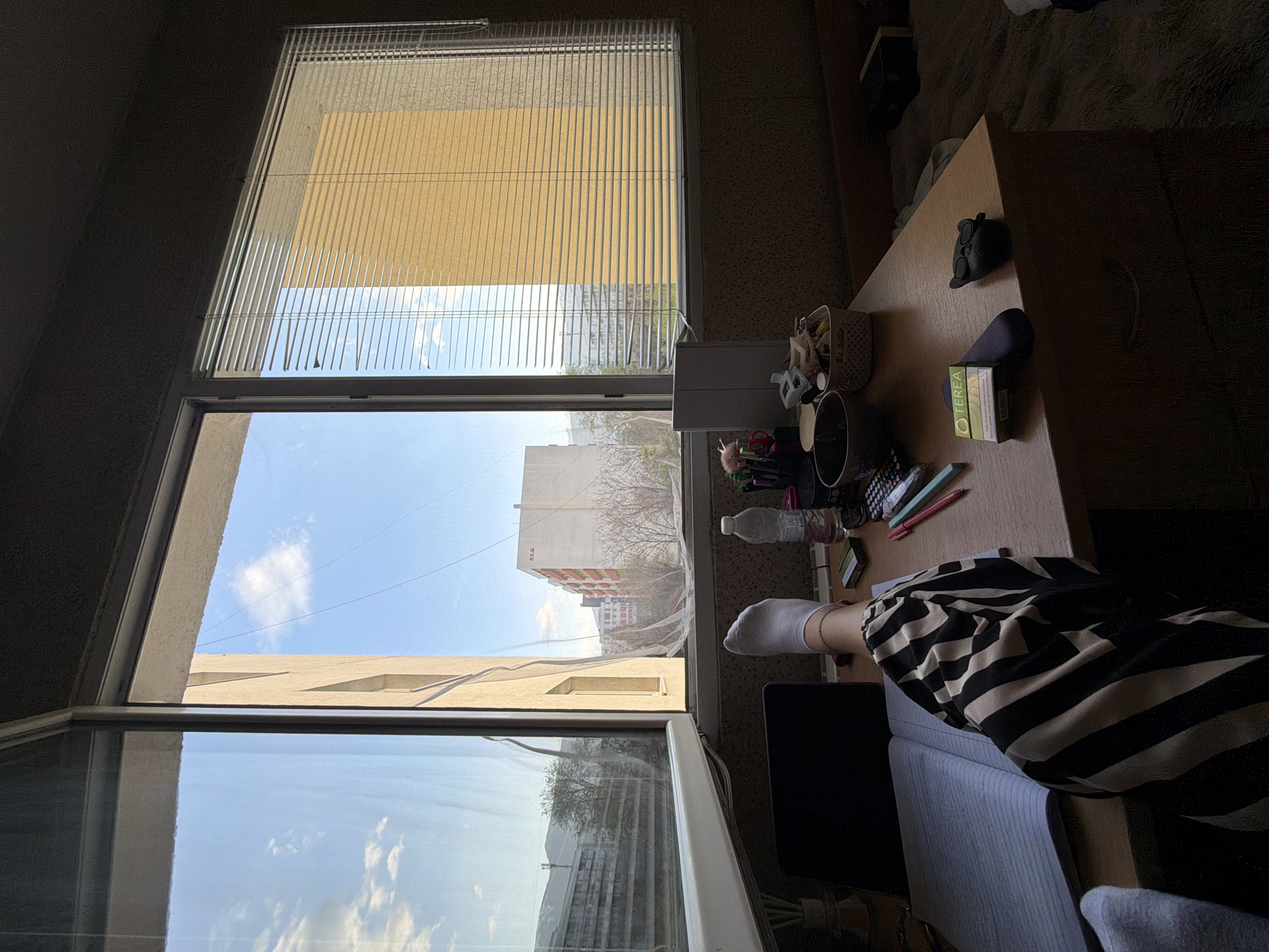 The image depicts an indoor scene, possibly an apartment, with a view through a large window overlooking a cityscape. A partially closed Venetian blind covers the top part of the window. Below the blind, the window reveals a clear blue sky and several buildings, including a white and red apartment building typical of Eastern European architecture. The buildings suggest an urban environment, potentially a residential area in Sofia, Bulgaria, given the architectural style and the overall context.
In the foreground, a wooden desk occupies a significant portion of the frame. The desk is cluttered with various items, including a basket, a cup holding writing utensils, a water bottle, pens, and a pack of cigarettes. A black object resembling eyewear or a small toy sits on the desk's surface.
A person is seated at the desk, with their legs visible. They are wearing a white sock and striped black and white pants. A light blue sofa or chair is partially visible beneath the person's legs. The image suggests someone is working or spending time at the desk while looking out of the window. The overall lighting indicates it is daytime.