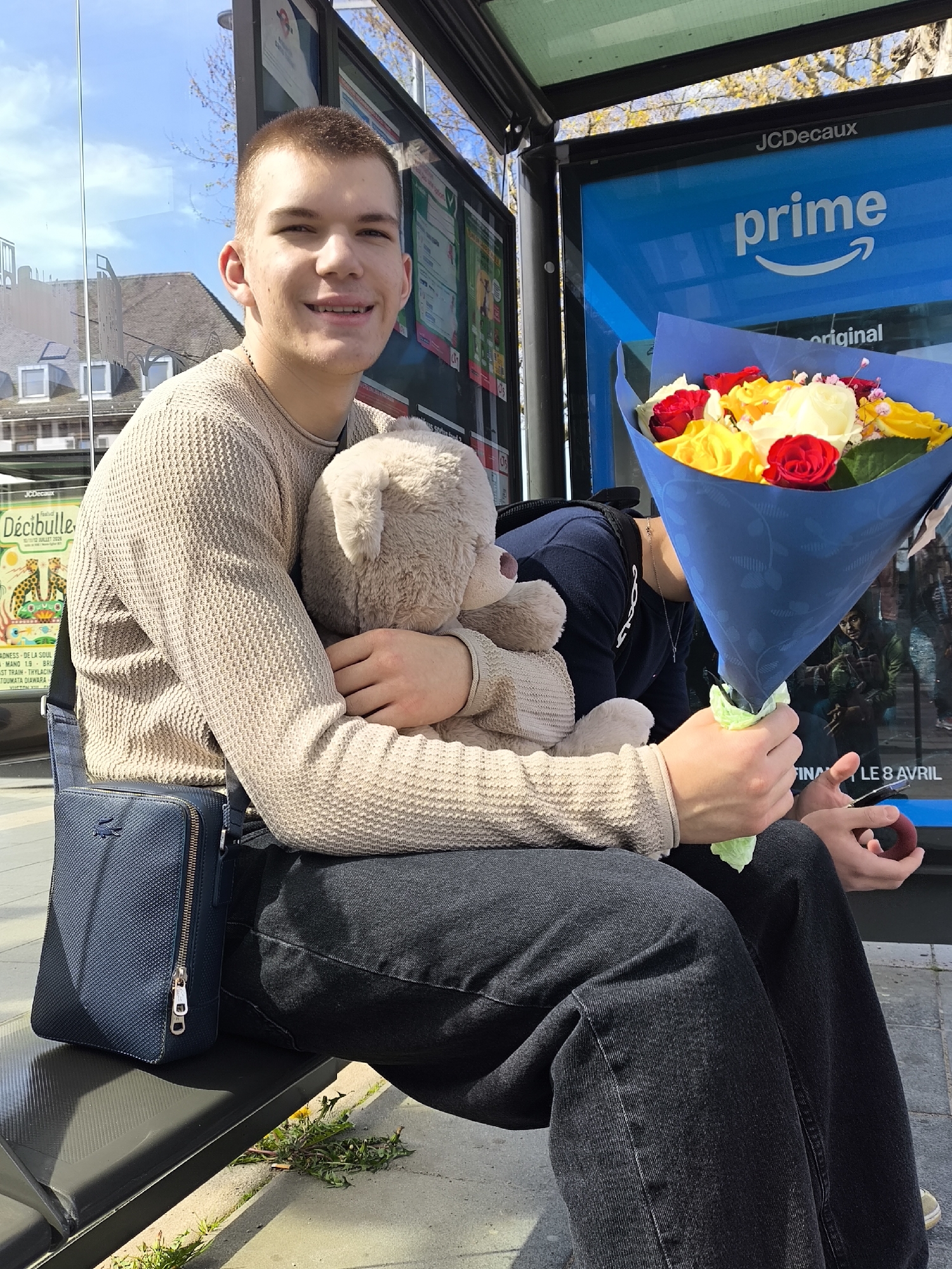 A young man sits at a bus stop in Strasbourg, France, embracing a teddy bear and holding a bouquet of red, yellow, and white roses. The bus stop has advertisements for JCDecaux, Prime, and the "Décibulle" festival. The man wears a beige knitted sweater, black jeans, and a dark blue Lacoste cross-body bag. The bus stop offers a view of buildings across the street.