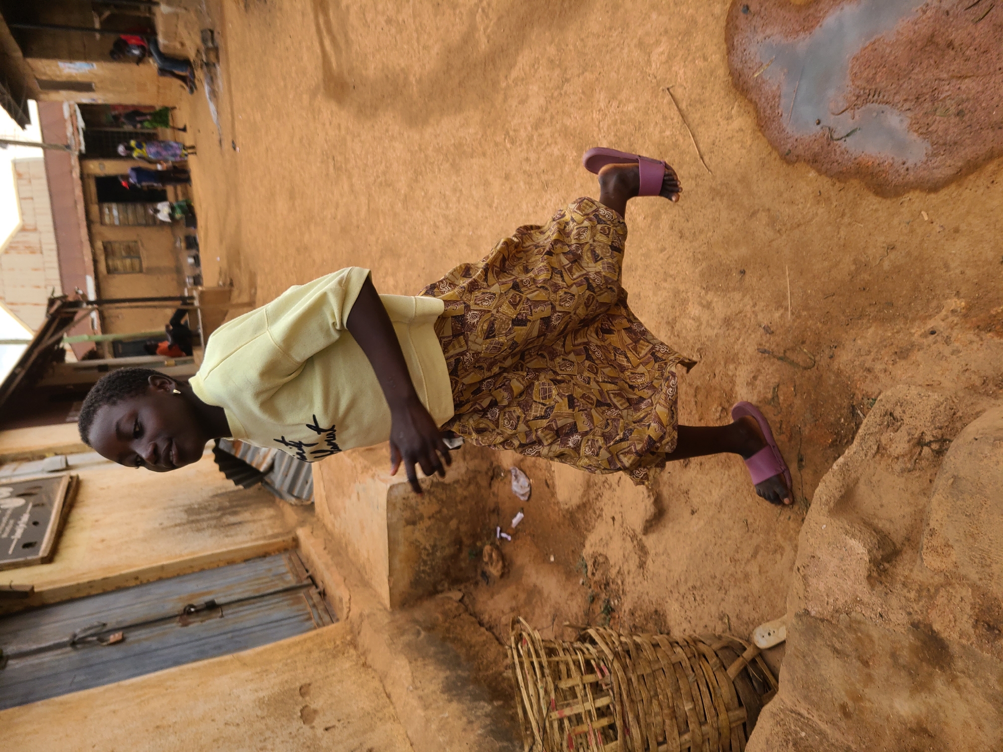 A young girl in Jos, Nigeria, is seen from a slightly elevated angle. She wears a light yellow t-shirt and a brown patterned skirt with pink sandals. She is standing on a dirt surface with a large puddle of water nearby. In the background, there are buildings with a few people walking in the distance. A woven basket and a corrugated metal sheet are visible in the foreground.