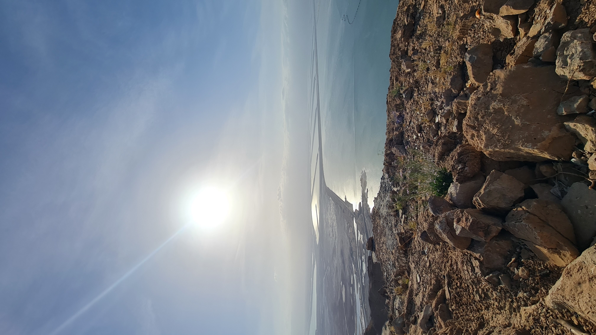 The image shows a landscape in the Tamar Regional Council, Israel, featuring a rocky cliff in the foreground. Beyond the cliff, there is a body of water, possibly the Dead Sea, reflecting sunlight. The sky is blue with some clouds, and the sun is bright. The water appears to have rectangular shapes in it.