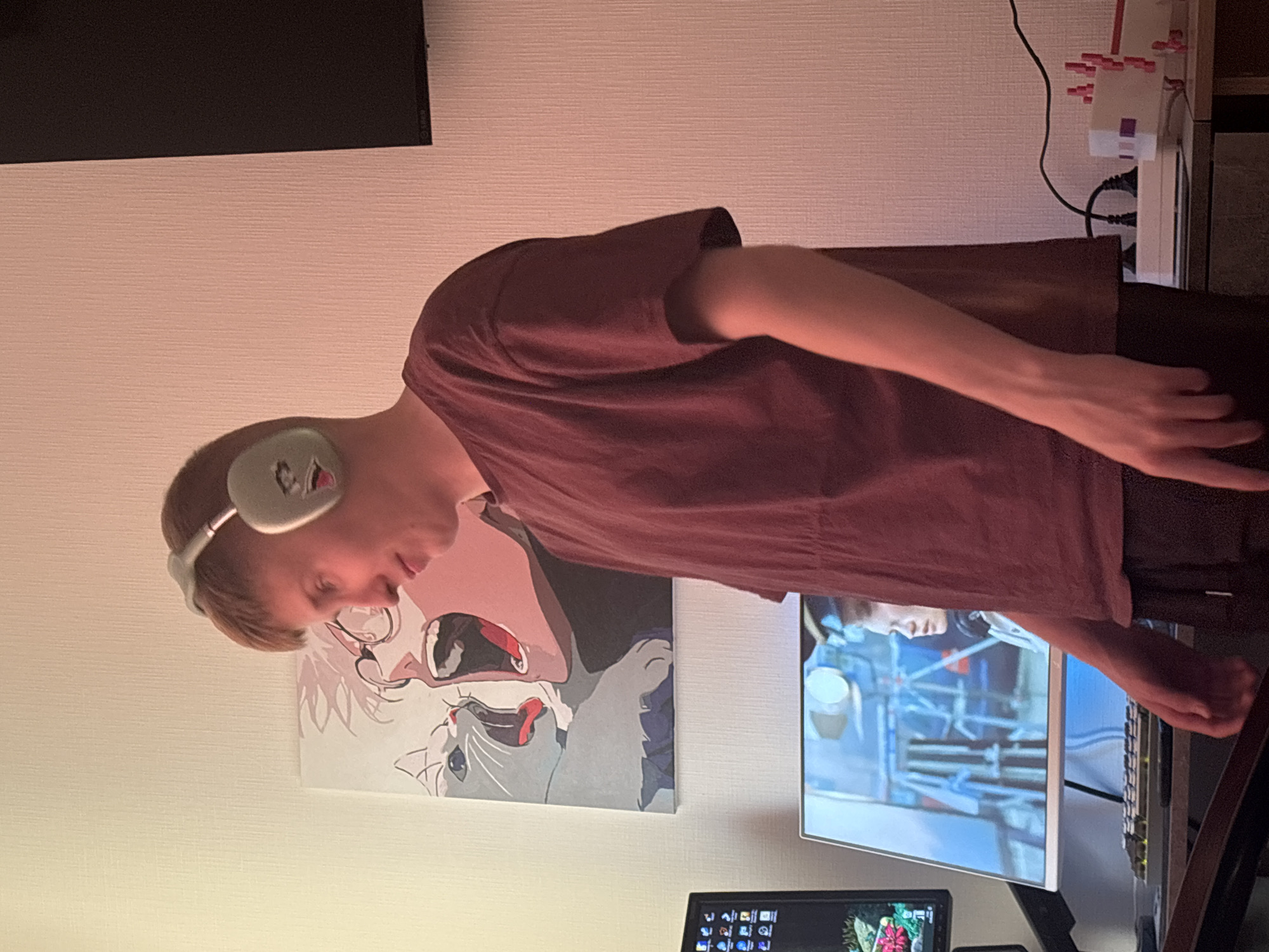 A young person with short, light-brown hair, wearing clear-framed glasses and light-colored over-ear headphones, is depicted in an indoor setting. They are dressed in a maroon short-sleeved t-shirt and are looking intently towards a computer monitor, their head tilted slightly forward. Their right arm and hand are visible, resting near a keyboard or mouse area, suggesting engagement with the computer.
The setting appears to be a personal room or study in Sinyavino, Russia, illuminated by artificial light, indicative of evening or nighttime. On the desk in front of the person, a large monitor displays a video or game scene featuring a person's profile and industrial-like structures. Below this, a smaller monitor shows a desktop with numerous icons and a vibrant, possibly floral, background image. A keyboard is visible on the desk.
On the wall behind and to the left of the person, an artwork depicts an anime-style face with glasses and an open mouth, alongside a white cat with an open mouth. To the right, a white power strip with multiple red plugs is mounted on the wall, connected by black cables. A television screen, with the "BENQ" logo faintly visible on its frame, is mounted high on the wall in the background. The walls feature a light-colored, textured finish. The overall ambiance suggests a focused activity, likely gaming, watching media, or working, centered around digital interaction.