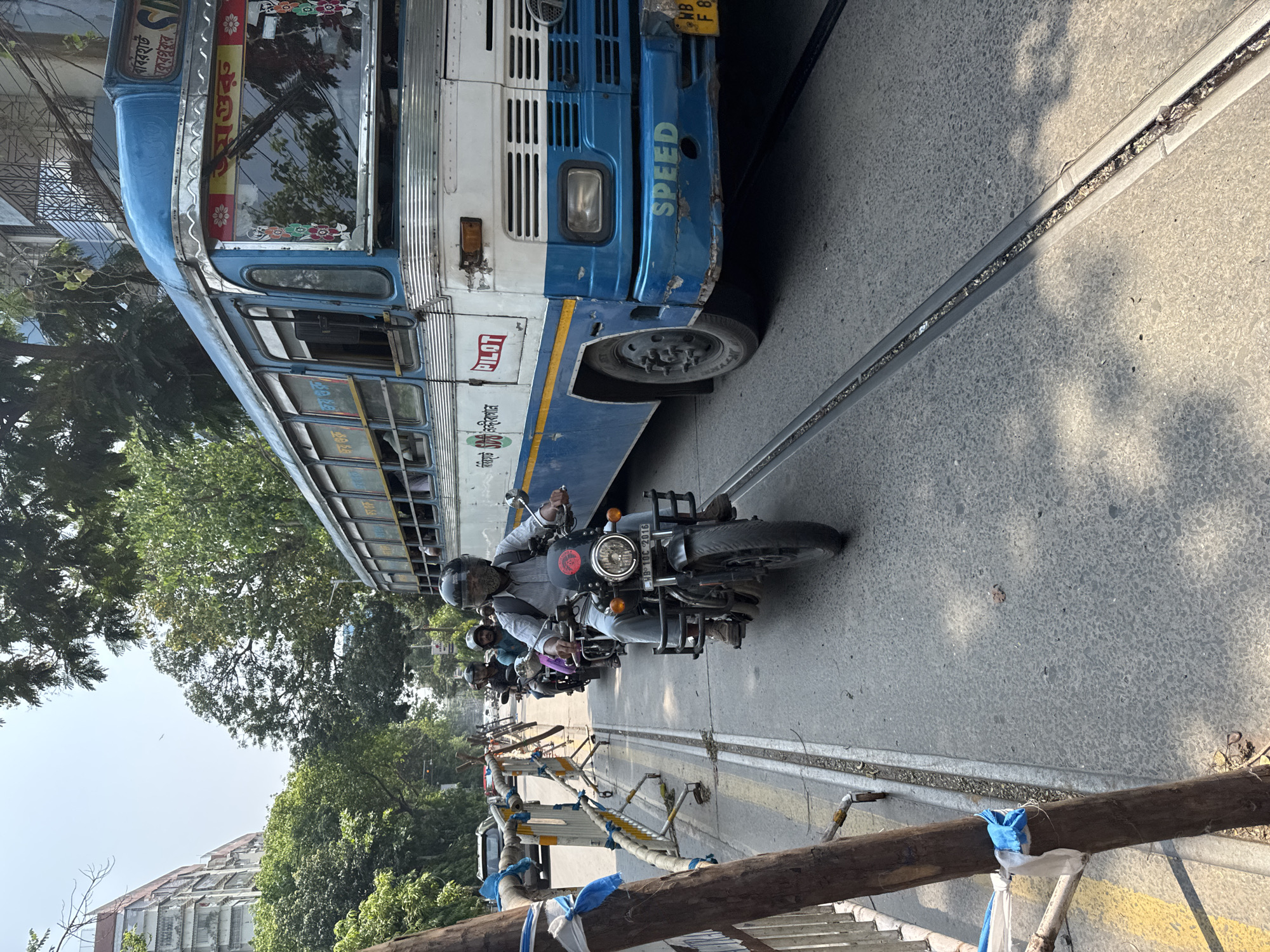 The image depicts a street scene in Kolkata, India. A blue and white public transit bus, with the word "SPEED" painted on its front, is partially visible on the right side of the frame, its front tire nearly aligned with a tram track embedded in the pavement. A man wearing a helmet and light-colored clothing is riding a black motorcycle next to the bus, on the tram tracks. Other motorcycles and riders follow behind him. Barriers made of wooden planks and metal frames are set up along the side of the road. Trees and buildings line the opposite side of the street. The sky is hazy, suggesting a clear but perhaps polluted atmosphere. The photograph appears to have been taken at street level.