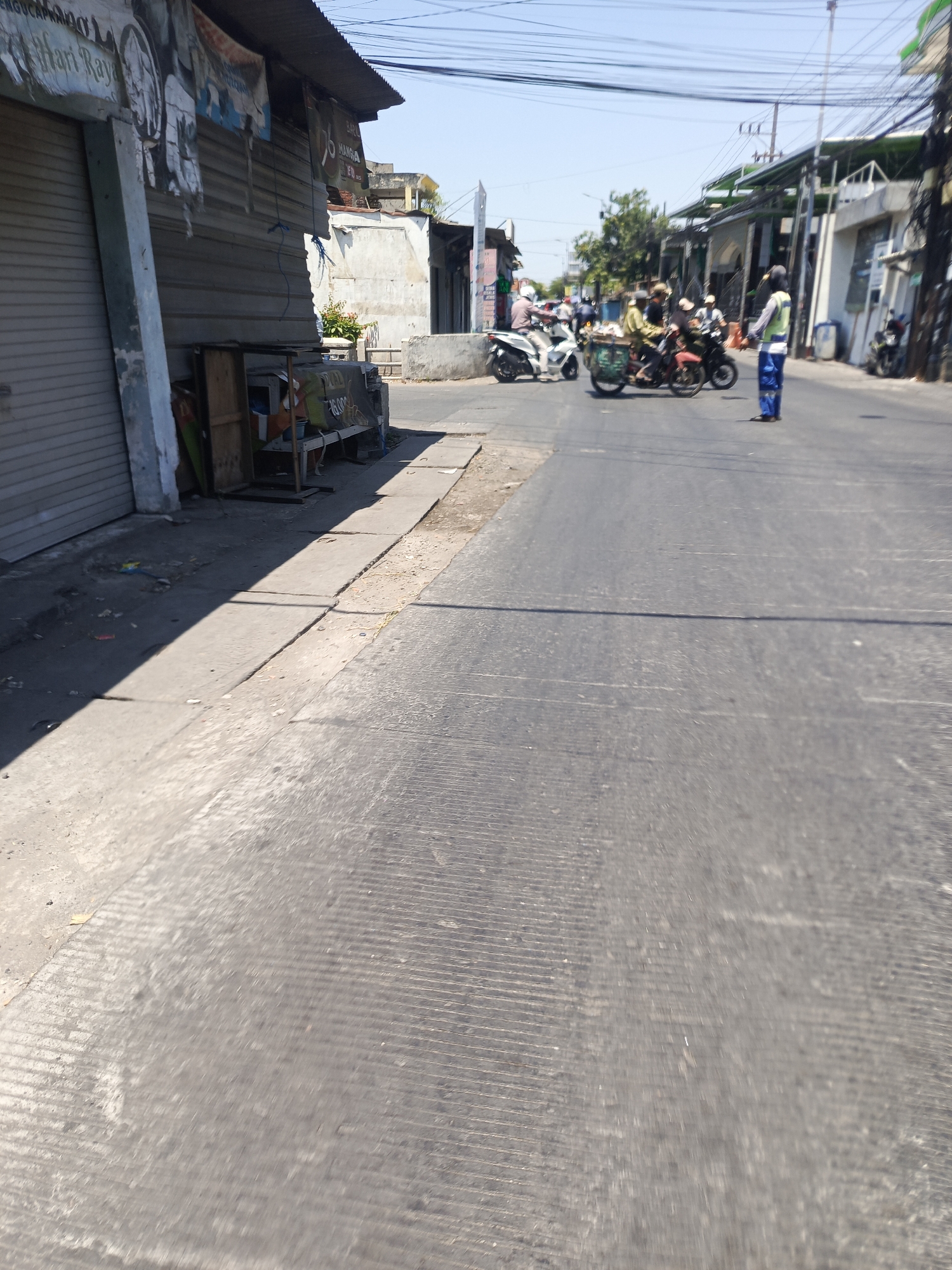 In the city of Kwangsan, Indonesia, a street scene unfolds under a clear blue sky. On the left, a row of shops with closed metal shutters lines the street. A faded banner hangs above one shutter. A small wooden cart is positioned near the entrance of one shop. A concrete sidewalk runs along the shops, casting a long shadow onto the road.
The street itself is paved with asphalt and shows signs of wear and tear, with prominent grooves running across its surface. Several people are on the road, including a person in a high-visibility vest and blue pants, appearing to be directing traffic. Three motorcycles are visible: one in the foreground carrying at least two people, and another further down the road with a rider. The background reveals more buildings and power lines, typical of an urban environment. The overall impression is a snapshot of daily life on a street in Kwangsan.