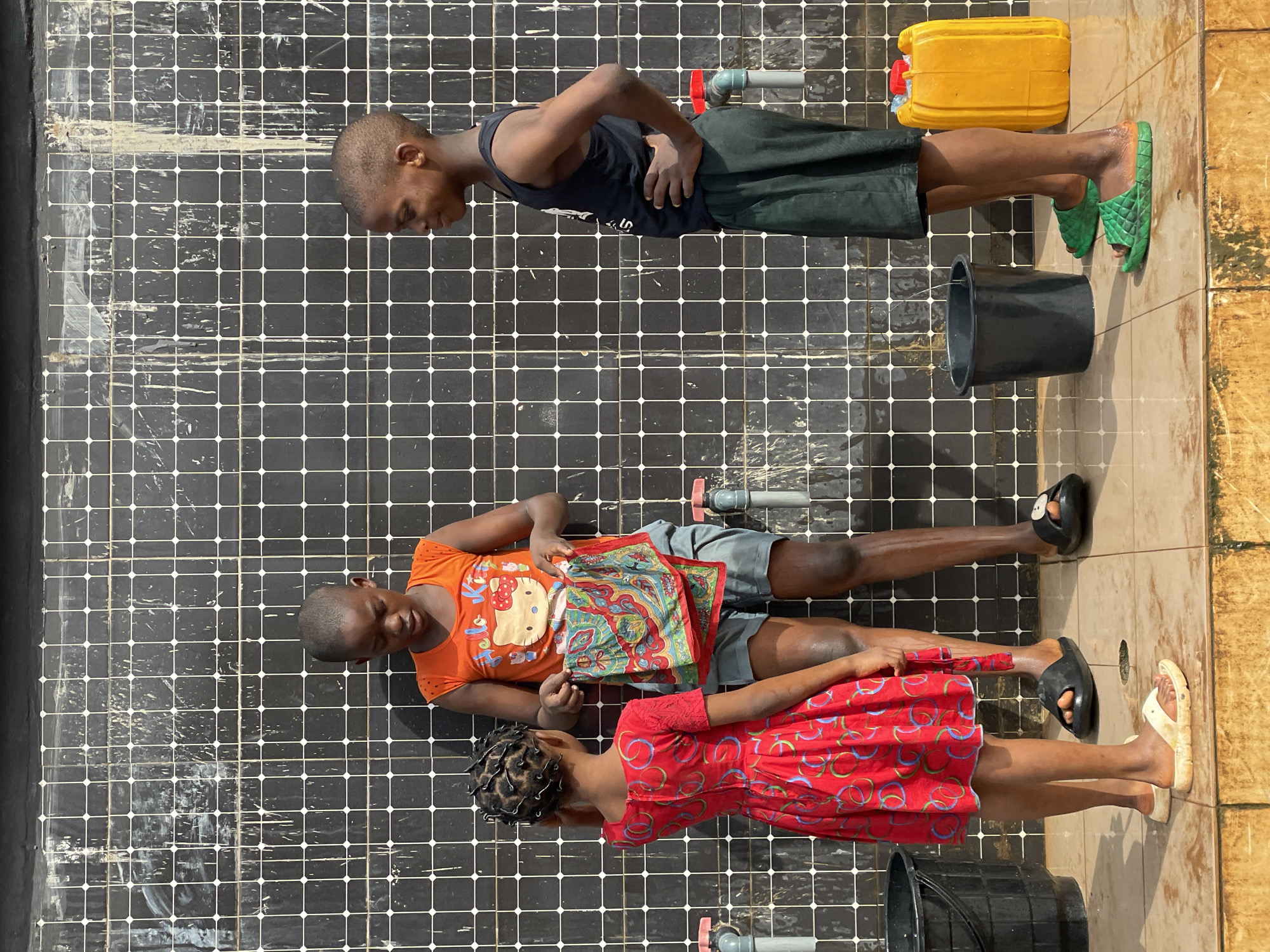 The image depicts three children outdoors in Nkpor, Nigeria. They stand on a tiled surface, possibly concrete, divided into small black squares with white grout lines. This surface appears to be wet and streaked. A young boy stands at the top, wearing a dark tank top and dark shorts, with green sandals. A yellow jerrycan and a black bucket are nearby. Below him are a second young boy and girl. The boy wears an orange t-shirt, gray shorts, and dark sandals. The girl, positioned at the bottom, wears a red patterned dress and light-colored sandals. A black bucket is near her, and a patterned cloth lies between them. A pipe with a red valve extends from the black tiled surface between each person. The scene is brightly lit with sunlight.