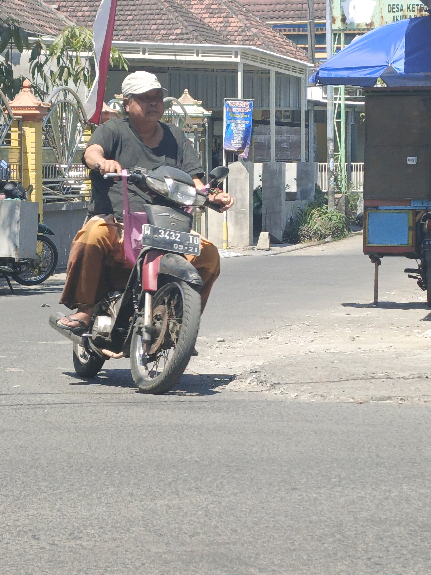 The image shows a man riding a motorcycle on a street in Ketegan, Indonesia. The man is wearing a dark t-shirt and a light-colored cap. He is also wearing loose, orange pants and flip-flops. The motorcycle is red and black and has a license plate that reads "W 3432 TQ 09-21". A pink bag is hanging from the handlebars. In the background, there are buildings with tiled roofs, a blue umbrella, and a street cart. The ground is paved with asphalt and gravel.