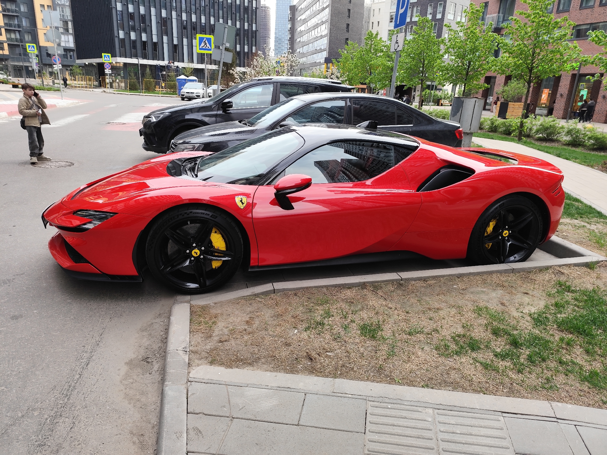 The image depicts a red Ferrari SF90 Stradale parked on a city street in Moscow, Russia. The car is parked parallel to the curb, with a black SUV parked behind it. To the left of the Ferrari, a young person dressed in a tan jacket, grey pants, and brown shoes stands on the sidewalk talking on a mobile phone. In the background, there are modern buildings, trees, street signs, and pedestrians. The street is paved with asphalt, and the sidewalk is made of concrete tiles. A strip of grass and dead vegetation is situated between the curb and the sidewalk.