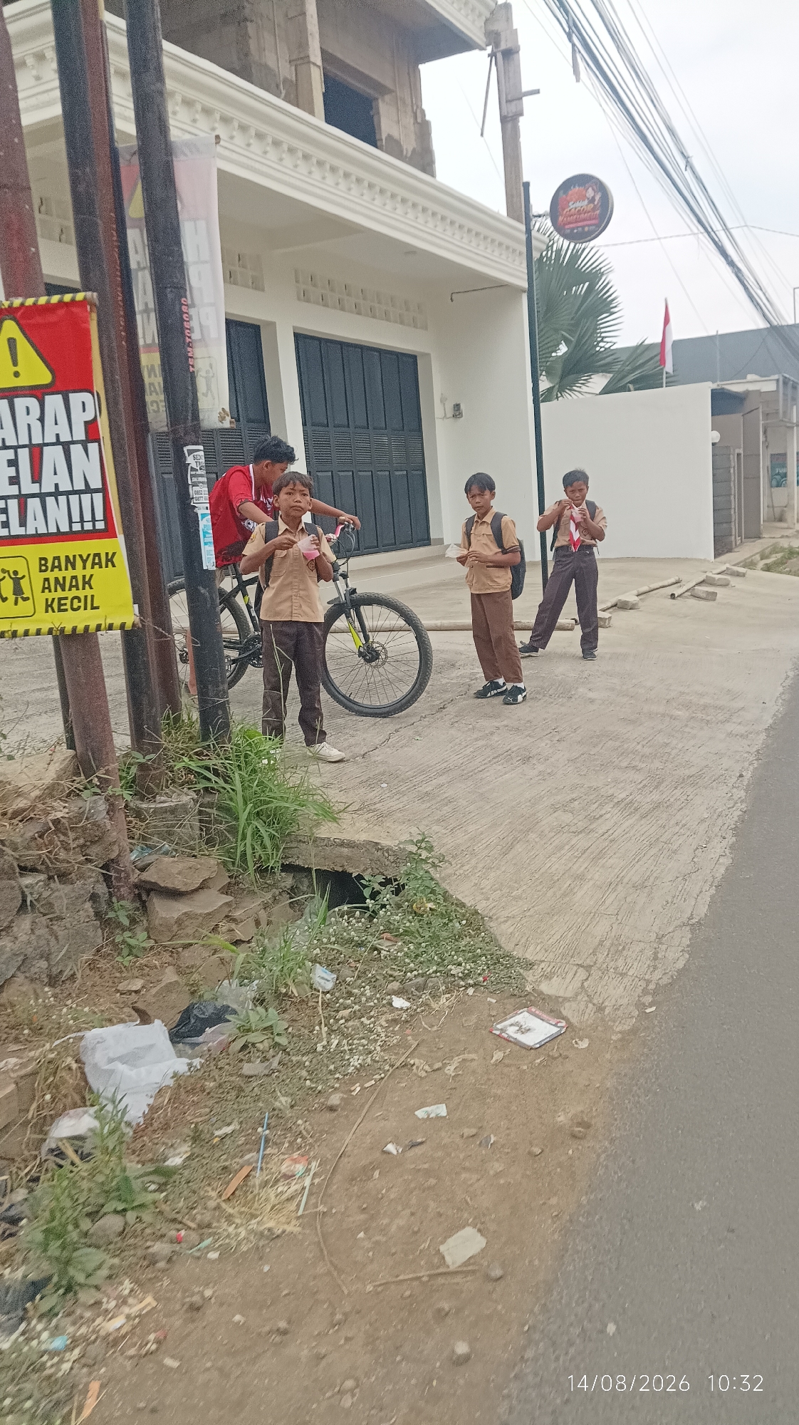 The image captures a street scene in Tasikmalaya, Indonesia, on a cloudy day. In the foreground, a paved road meets a dirt and gravel area with visible litter, including plastic bags and debris. Several young boys in school uniforms, consisting of light brown shirts and darker brown pants, are present. One boy in the front left is holding a pink cup. Another boy is standing next to a bicycle, and two others are further to the right, one of whom is holding a white object. A red shirt is visible behind the bicycle. In the background, a white building with dark-colored doors and windows is under construction. A sign with yellow and red markings is attached to a pole on the left, and a round sign with an image and text is also visible on a pole. The overall environment suggests a semi-urban or suburban setting with elements of everyday life and ongoing development.