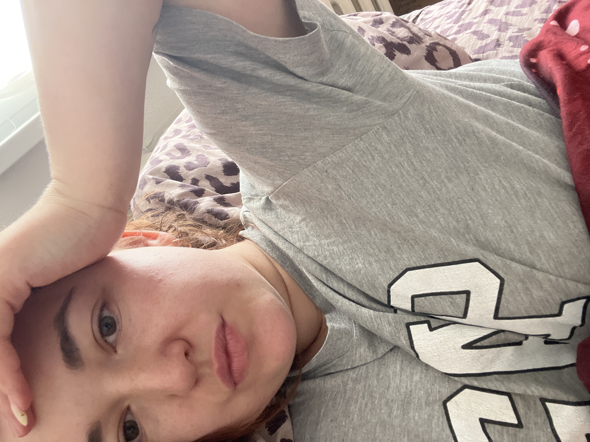 The image shows a close-up of a person's face and upper body, who is lying down. The person has pale skin, brown eyes, and reddish-brown hair. They are wearing a grey t-shirt with the word "GAP" printed on it in white letters with a black outline. Their right hand is raised and resting on their forehead. The background consists of bedding with a leopard print pattern. There is no information in the image to indicate the location in the city of Chelyabinsk, Russia.