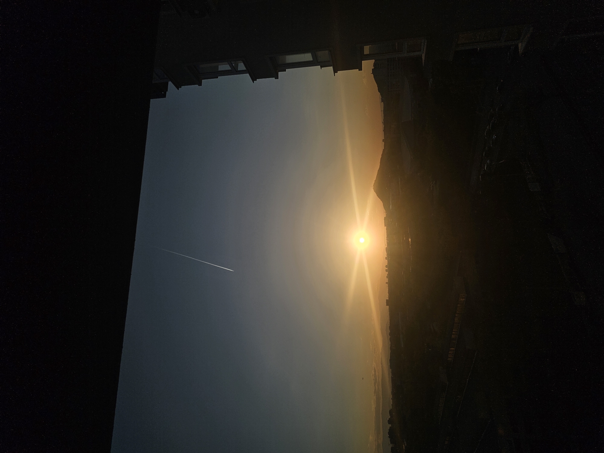 The image captures a serene sunrise or sunset view over Plovdiv, Bulgaria, seen from a building, likely a window or balcony. The scene is dominated by a brilliant sun, positioned towards the right side of the frame, emitting strong golden light and a starburst effect. The sky transitions from a deep, clear blue-grey on the far left to vibrant orange and gold hues closer to the sun, indicating either dawn or dusk.
Across the upper left portion of the sky, a faint, white contrail left by an airplane is visible, extending diagonally upwards.
In the foreground, the dark silhouettes of buildings frame the top and bottom edges of the image, suggesting an urban setting. Their distinct shapes, including windows and structural elements, are discernible against the brighter sky.
Along the horizon, stretching from beneath the sun and across the mid-right portion of the image, are the dark, indistinct silhouettes of mountains or hills, blending into a hazy landscape of a city. The strong backlighting from the sun renders these distant features in deep shadow.
The overall impression is one of peaceful solitude, with the dramatic natural light creating a strong contrast between the silhouetted foreground and the luminous sky. No people or visible text are present in the image.