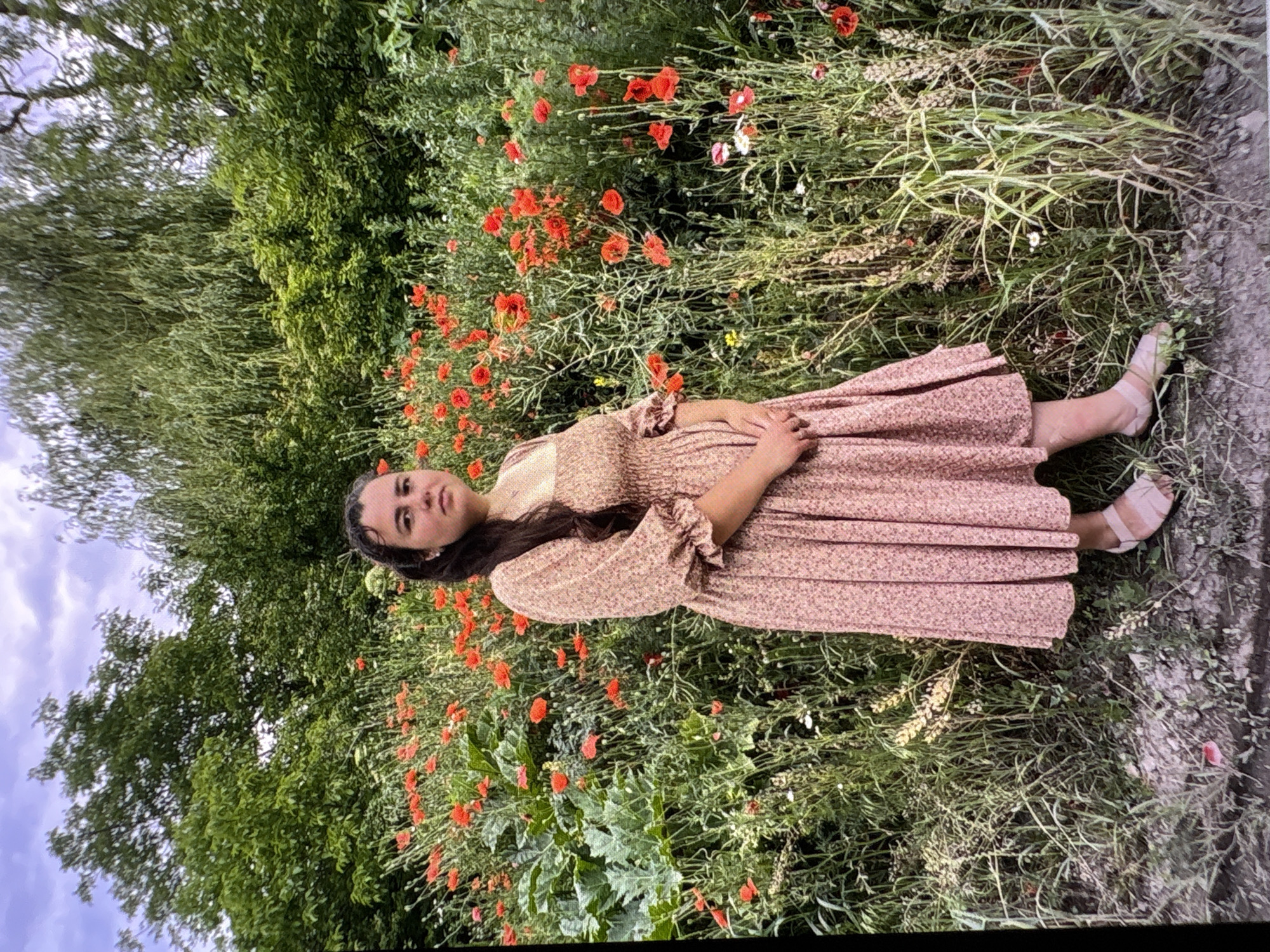 A full shot shows a young woman standing in a field of red poppies. She is wearing a pink floral dress with puffed sleeves and a square neckline. Her hands are clasped in front of her. The woman has dark hair pulled back in a ponytail and is looking directly at the camera. The background consists of lush green trees and a cloudy sky. The ground is covered in grass and dirt, with a few scattered pebbles. The scene is outdoors and appears to be taken during the daytime.