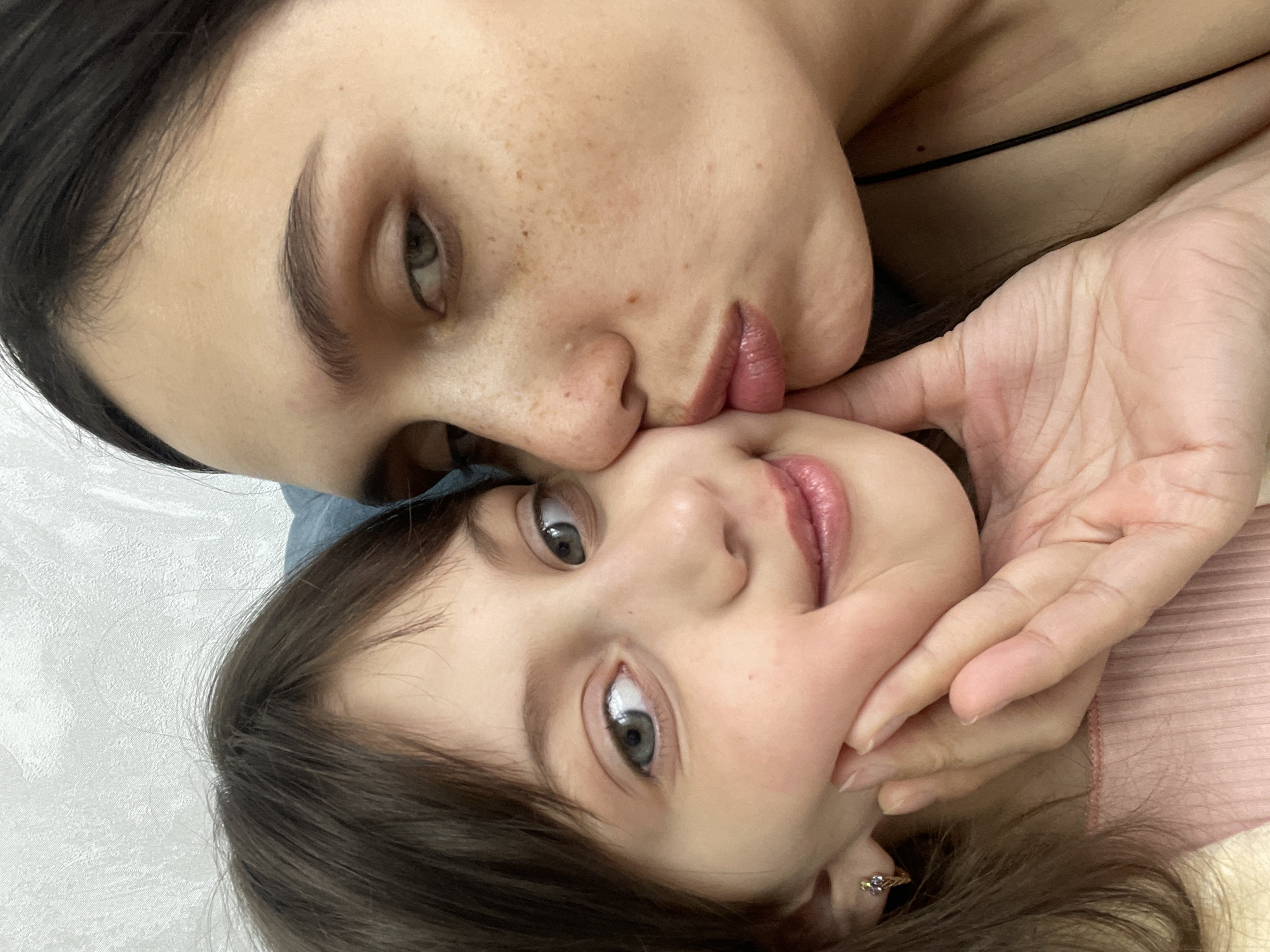 A selfie shows a woman with dark hair and fair skin kissing a child, presumably a daughter, on the cheek. The woman has green eyes and freckles. The child has dark hair and light blue eyes. A hand is placed near the child's face. The background is out of focus and does not provide sufficient information to determine the location in Izhevsk, Russia.