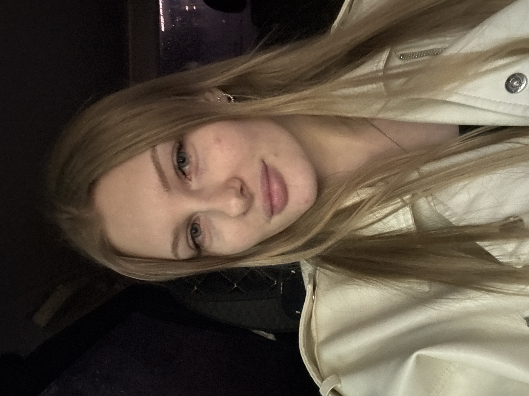 A close-up, slightly angled selfie of a young woman with fair skin and long blonde hair wearing a light-colored jacket. The setting appears to be indoors, possibly a dimly lit room. The lower left corner of the image shows dark fabric that might be clothing or furniture. The woman's face is the primary focus. It is not possible to determine a specific location within Perm, Russia, from the image.
