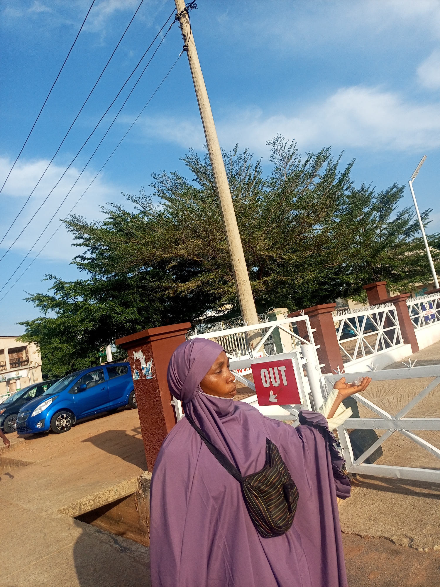 The image presents an outdoor scene, likely a street or compound entrance in Gombe, Nigeria, under a bright blue sky with sparse white clouds. In the foreground, partially cropped on the right, a person is visible from the waist up, dressed in a flowing purple abaya and hijab. They wear a striped black and multi-colored shoulder bag. Their left hand is raised and open, holding a small, translucent object, possibly receiving or gesturing. The person's face is seen in profile, looking slightly upwards and to the right.
To the right of the person, a white metal gate or fence is prominent, featuring a red rectangular sign that clearly displays the word "OUT" in white capital letters, accompanied by a small downward-pointing hand icon. A brown pillar stands next to the gate. In the middle ground, a vibrant blue car, possibly a Chevrolet Spark, is parked on a paved surface, with another darker car partially visible behind it. The ground directly in front of the person is uneven, appearing to be a mix of concrete and unpaved earth, with distinct shadows indicating bright sunlight.
A tall wooden utility pole with multiple electrical wires extends diagonally across the upper left portion of the frame, contrasting against the sky. A dense canopy of lush green trees forms the background, extending across much of the mid-ground. Distant buildings are also visible on the far left. The scene depicts a clear, sunny day in what appears to be an urban or semi-urban environment.
