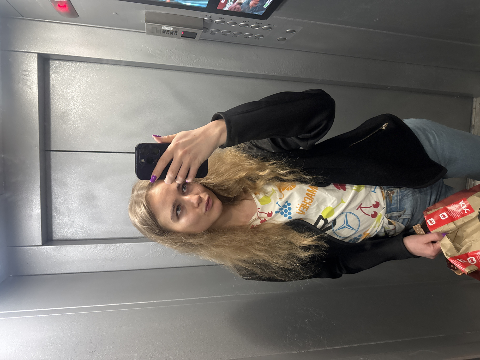 A person is taking a selfie in an elevator. The person has long blonde hair and is wearing a black jacket over a white t-shirt with a Mercedes Benz logo and fruit designs. They are holding a black smartphone with purple nail polish on their fingers. In their other hand, they are holding a brown paper bag from KFC. The elevator has metal doors, a control panel with buttons and a digital display, and a small video screen at the top. The location is in the city of Unknown, Unknown.