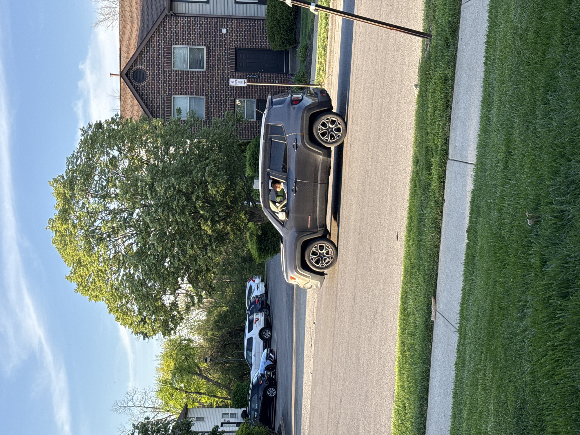 A slightly elevated daytime shot of a residential street in Columbus, Ohio. A dark grey Jeep Renegade is parked parallel on the right side of the street, facing the viewer. The driver is visible inside the car. Behind it, a row of other parked cars, varying in color (white, dark blue, grey), stretches further down the street. The right side of the road has a sidewalk bordering a lush green lawn. On the left, a large tree obscures part of a multi-story brick building. The street has a "No Parking" sign affixed to the front of the building, indicating parking regulations. The sky above the tree is blue with sparse white clouds.