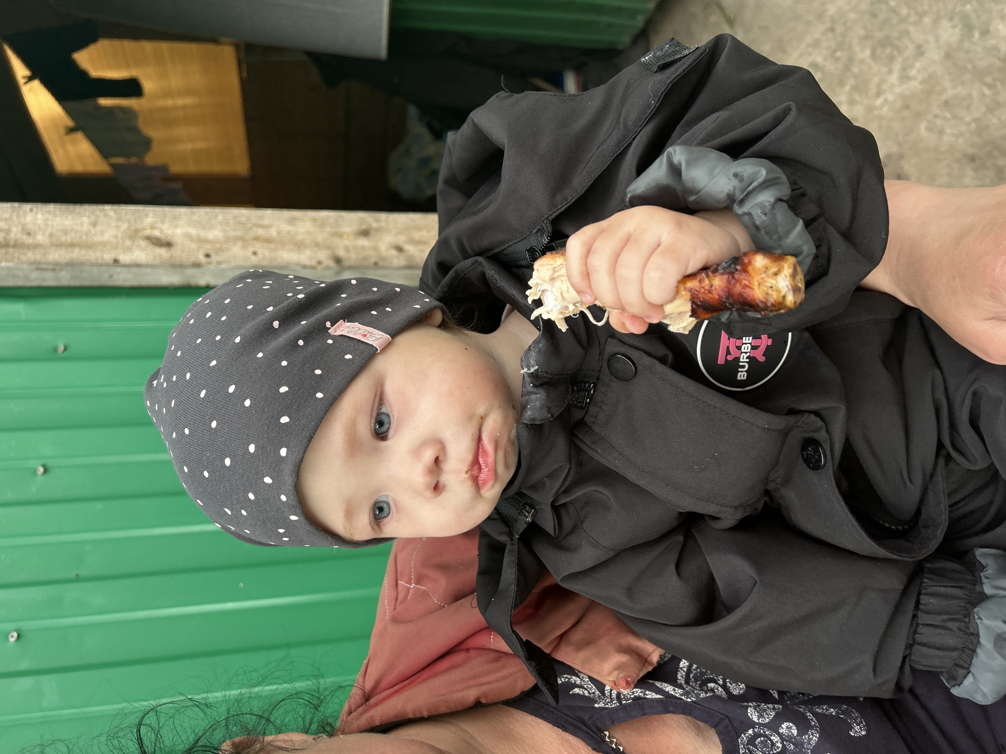 A young child with blonde hair and blue eyes, wearing a grey polka-dot beanie and a black jacket, is being held by an adult. The child is eating a piece of food. The background shows a green corrugated metal wall, a wooden beam, and laundry hanging on a line in the distance, suggesting an outdoor, possibly rural or informal, setting. The location is identified as Азинский, Russia.