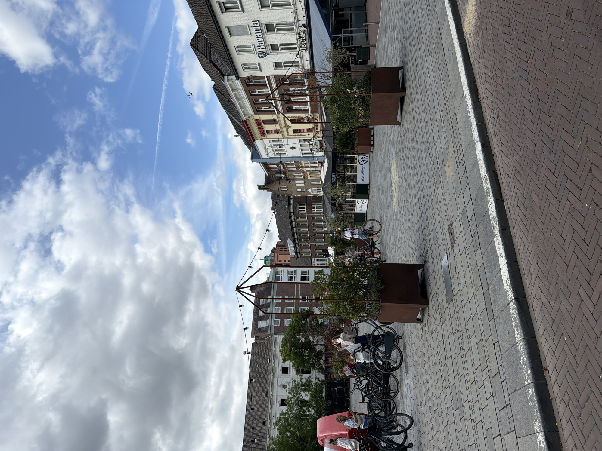 The image depicts a street scene in Roermond, Netherlands. In the foreground, the ground is paved with interlocking grey stone bricks, with a distinct curb lined with white paint. To the right, a herringbone pattern of dark red bricks is visible. In the middle ground, several people are gathered around bicycles. A group of four women, some of whom are holding phones, stand near a red, inflatable-like structure. To the right of the women, a row of parked bicycles is visible. Further back, in the background, a row of European-style buildings line the street. One building prominently displays the "Bavaria" logo. String lights are strung across the street between poles. Two large, square planters with greenery are placed on the paved area. The sky above is partly cloudy, with patches of blue sky visible.