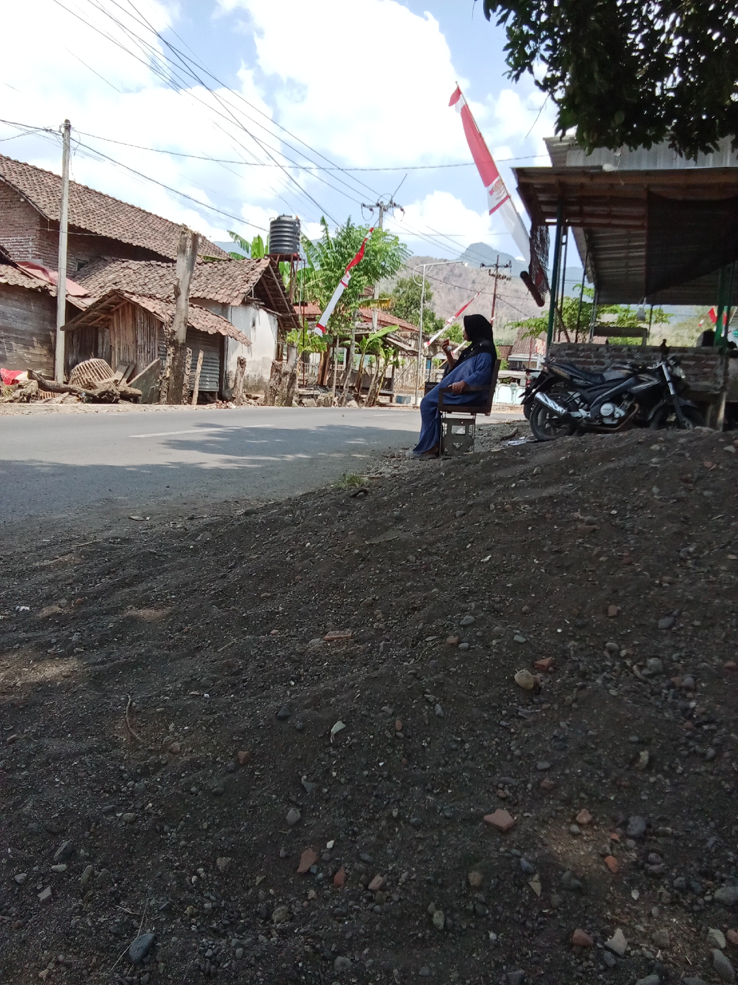 A woman wearing a black hijab and blue clothing sits on a metal chair next to a dirt pile, looking towards the left. In the background, a road runs horizontally across the image, bordered by simple buildings with tiled roofs and wooden structures. A black motorcycle is parked to the right of the woman. Red and white Indonesian flags are visible, one flying from a pole and another draped from a canopy structure. Power lines crisscross the sky, and distant hills are visible under a cloudy blue sky. The foreground consists of a rough dirt and gravel surface. The scene appears to be in a rural or semi-rural area.