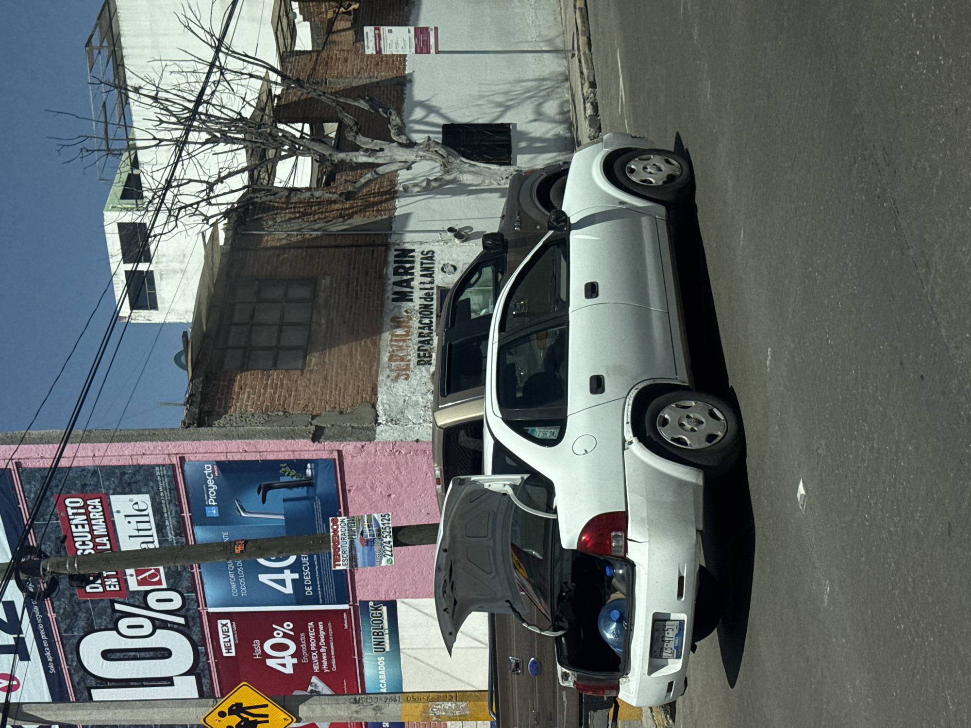 The image captures a daytime scene in Puebla, Mexico, depicting an urban street with buildings and a parked car. The main subject is a white two-door car, possibly a Chevrolet Corsa, parked on the right side of the road with its trunk open, revealing two blue plastic water bottles inside. Behind it, part of a darker vehicle is visible.
The scene is dominated by a brick building on the left, which has "SERVICIO MARIN REPARACION DE LLANTAS" (Marin Service Tire Repair) painted on its facade, indicating a tire repair business. Above this building, a bare-branched tree stands in front of a white building. On the far left, a utility pole with multiple wires runs vertically, partially obscuring large billboards attached to a pink-walled building.
Notable details include:
- Bright sunlight and clear sky, suggesting good weather.
- The car's rear license plate reads "TXG-47-51".
- A small white sticker on the car's rear window says "TWG 95".
- The billboards display various advertisements in Spanish:
- "DESCUENTO EN LA MARCA altile" (Discount on the Altile brand)
- "Proyecta" (Project)
- "Hasta 40%" (Up to 40%) on "HELVEX, PROYECTA y Helvex By Designers" products.
- "ACABADOS UNIBLOCK" (Uniblock Finishes)
- "TERRENOS ESCRITURACIÓN PROPIEDAD VALSEQUILLO 2224-525125" (Land Property Deeds Valsequillo, with a phone number).
- A yellow cautionary road sign with figures is visible at the bottom.
The activity appears to be mundane street parking, possibly with the owner retrieving items from the car's trunk. The setting is characteristic of a bustling urban environment.