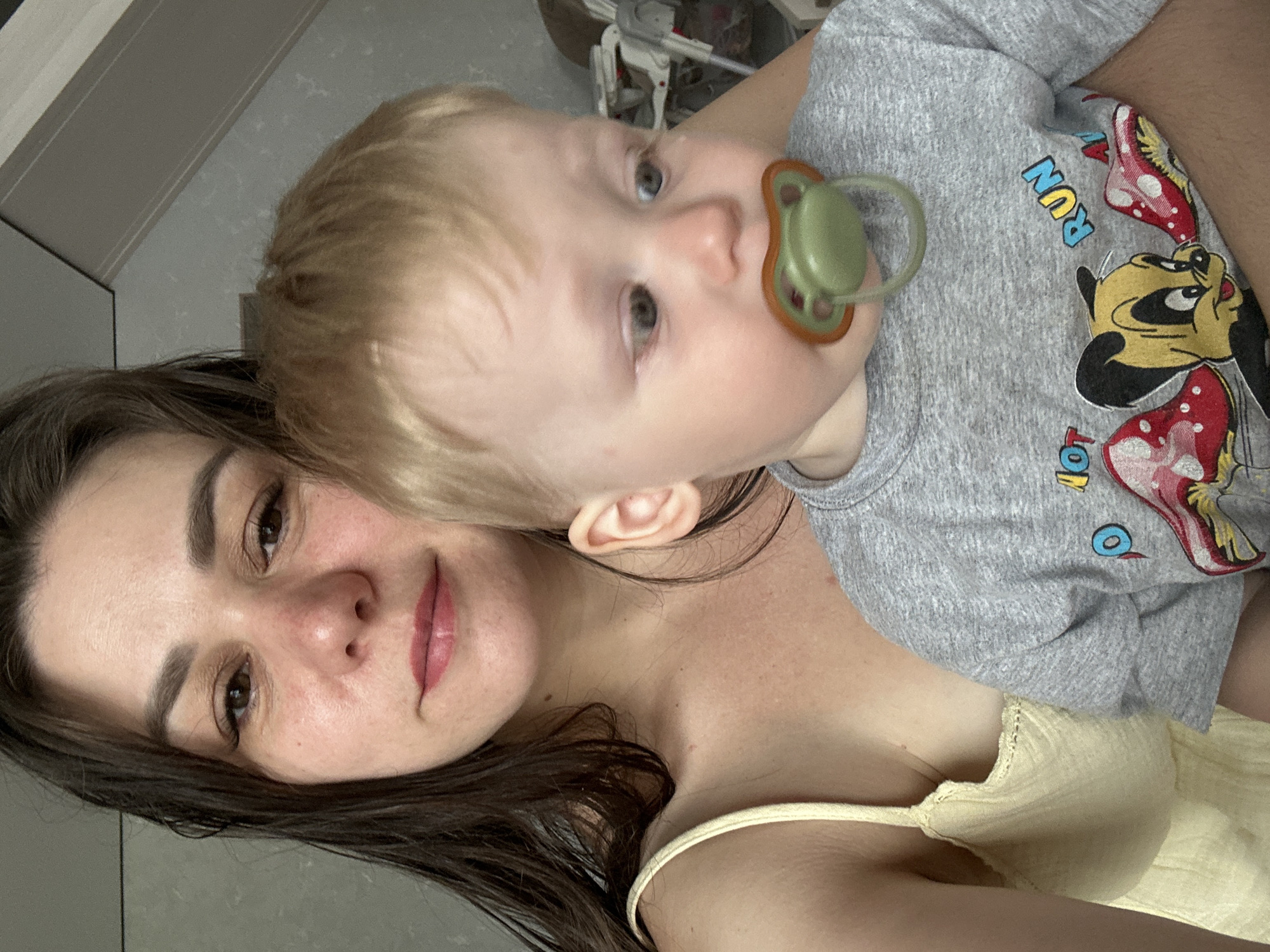 A woman with dark hair is holding a young child with blond hair in her arms. The child has a green pacifier in their mouth and is wearing a gray shirt with a Mickey Mouse design. The woman is wearing a yellow top. The location is unknown and there is no indication it is in Chaikovsky, Russia.