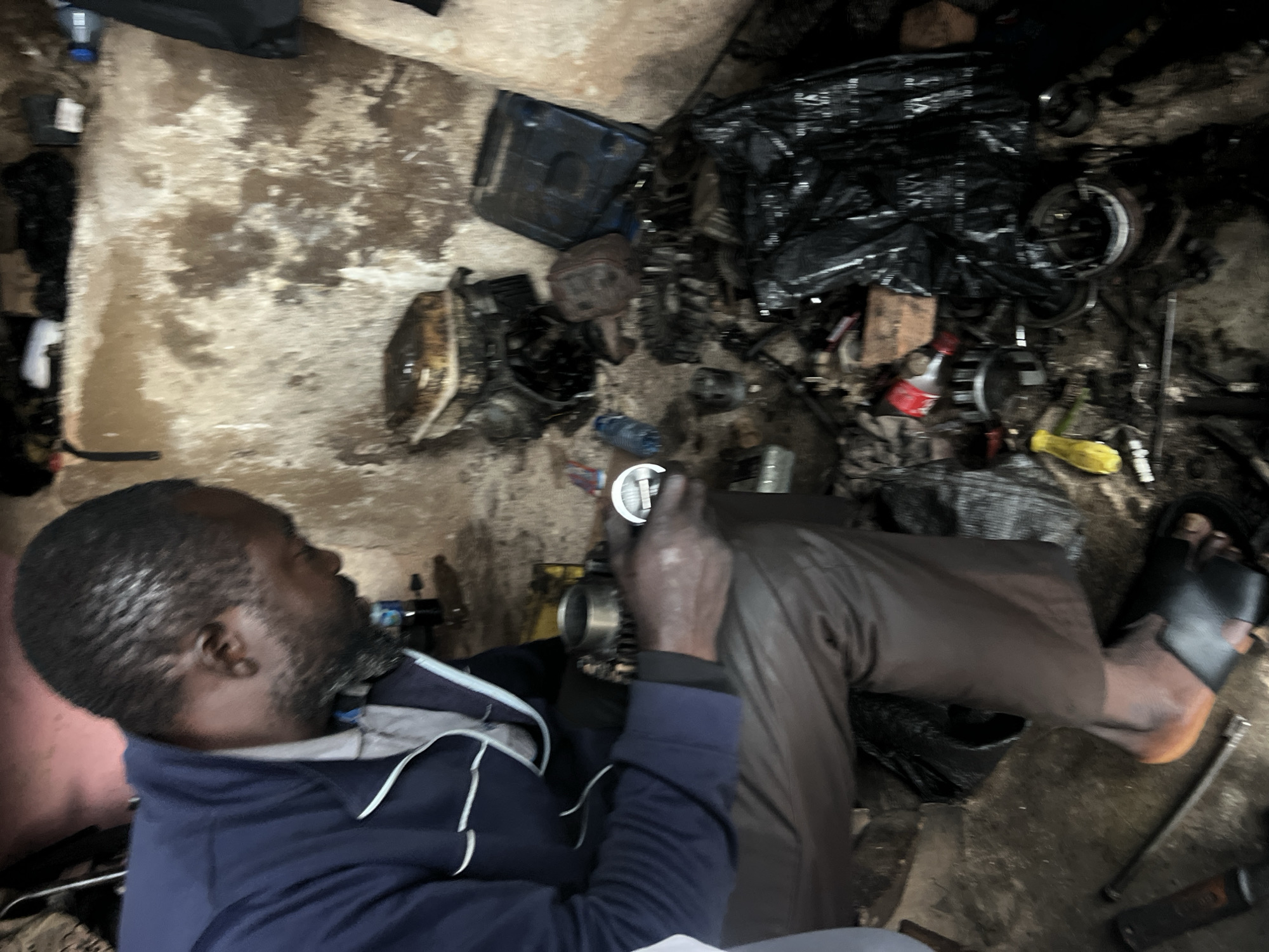 A man, identified as a mechanic, is seen working in a cluttered workshop located in Jos, Nigeria. He is seated, wearing a blue jacket over a light-colored shirt and brown pants. His attention is focused on the disassembled parts of what appears to be a small engine, which he is holding in his hands. The workshop environment is dimly lit, with rough, textured walls. Various engine components, tools, and miscellaneous items are scattered across the floor and surfaces, including a black plastic bag filled with parts, a rusty engine block, and a blue plastic bottle. The scene suggests an active repair or dismantling process.