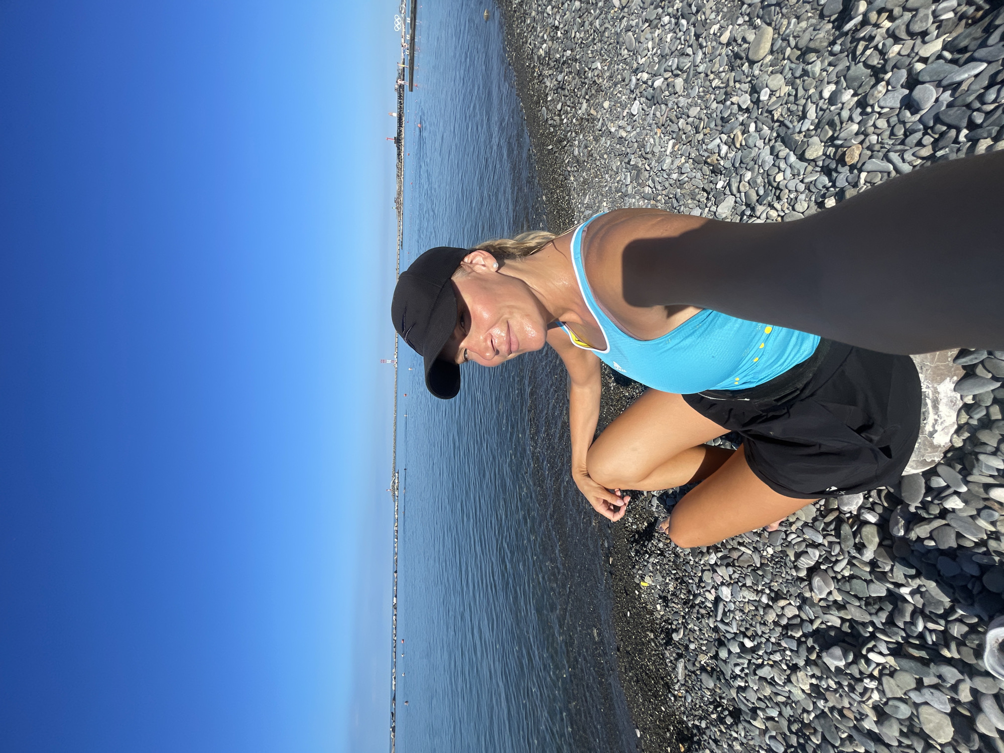 A woman wearing a blue tank top, black shorts, and a black baseball cap sits on a pebble beach facing the sea in Sochi, Russia. The sea is calm and blue, with a clear sky overhead. In the background, a long breakwater extends into the sea, with two lighthouses visible at its end. The beach is covered in smooth, grey pebbles. The woman is holding a phone in her right hand, appearing to take a selfie. Her left hand rests on her knee. The lighting suggests it is daytime, with the sun casting shadows on the beach.