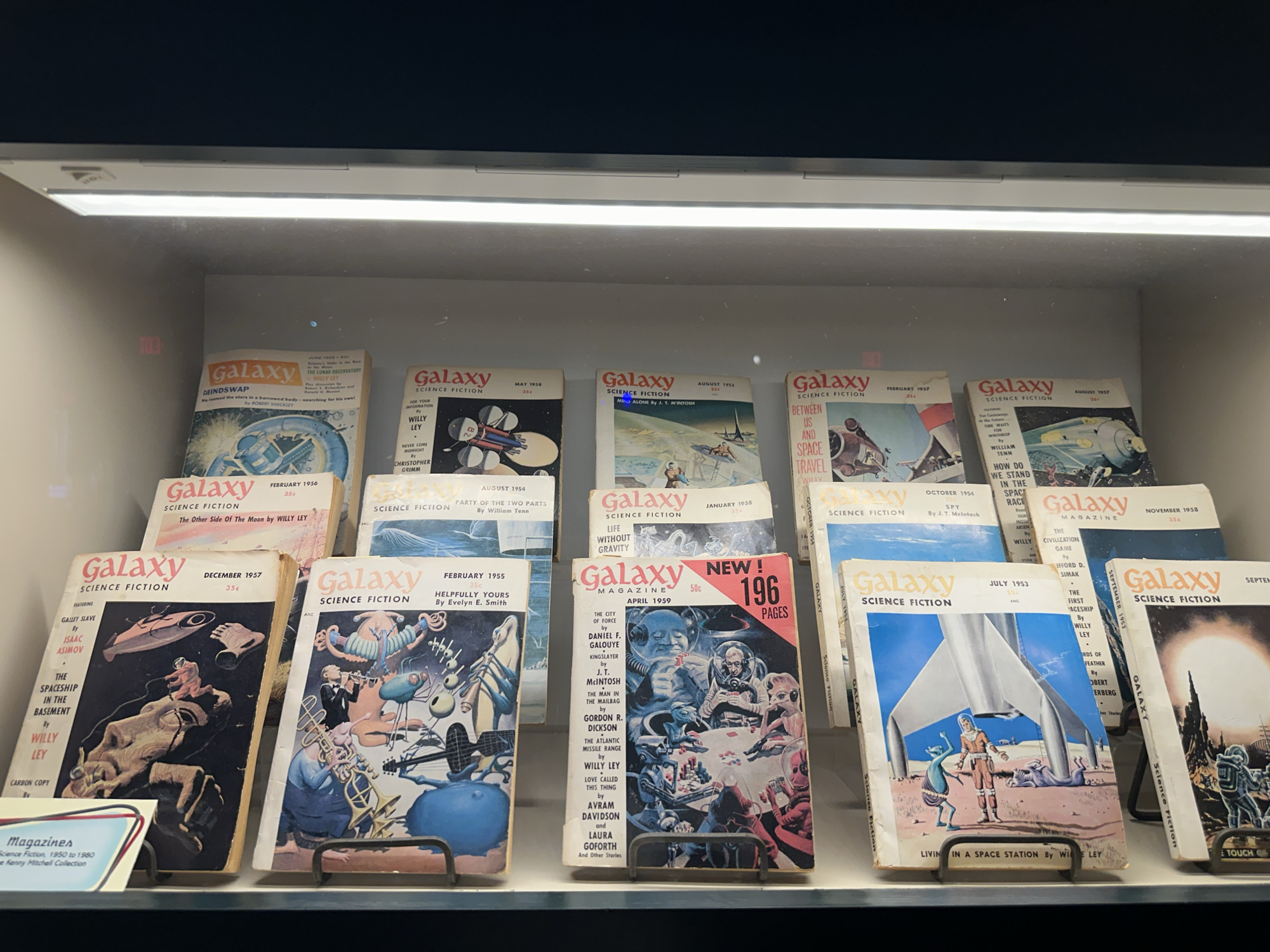 The image depicts a display case filled with vintage science fiction magazines, primarily from the publication "Galaxy Science Fiction." The magazines are arranged in stacks and rows on shelves. The titles and dates visible on the covers indicate a collection spanning several years, with prominent issues including "Galaxy Science Fiction" from December 1957, featuring a spaceship and a figure in a spacesuit; "Galaxy Science Fiction" from February 1956, with an illustration of the moon; and "Galaxy Magazine" from April 1959, with a cover depicting astronauts and aliens playing cards. Other visible dates range from 1953 to 1958. A small placard in the lower left corner identifies the collection as "Magazines, Science Fiction, 1950 to 1980, The Kenny Mitchell Collection." The display case appears to be illuminated by overhead lighting. The background is dark, suggesting the magazines are housed in a museum or exhibition setting. The overall impression is that of a curated collection of historical science fiction literature. This image is likely from the U.S. Space & Rocket Center in Huntsville, Alabama, given its extensive collection related to space exploration and science fiction.