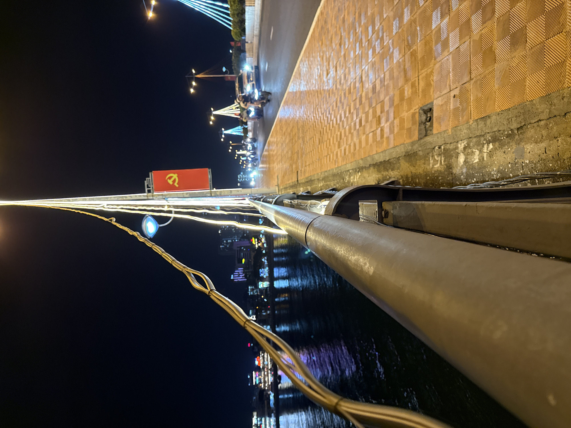 The image shows a nighttime view from a bridge in Nha Trang, Vietnam. On the left, a body of water reflects the lights of buildings and boats. A large metal railing runs along the foreground, with thick cables attached to it. In the background, a red flag with a yellow hammer and sickle is displayed on a tall pole, illuminated by white lights. The bridge has a tiled walkway and a road with traffic, including motorcycles and a car. Streetlights and decorative lights illuminate the scene. The overall atmosphere is that of a bustling city at night, with reflections on the water and illuminated architecture.
