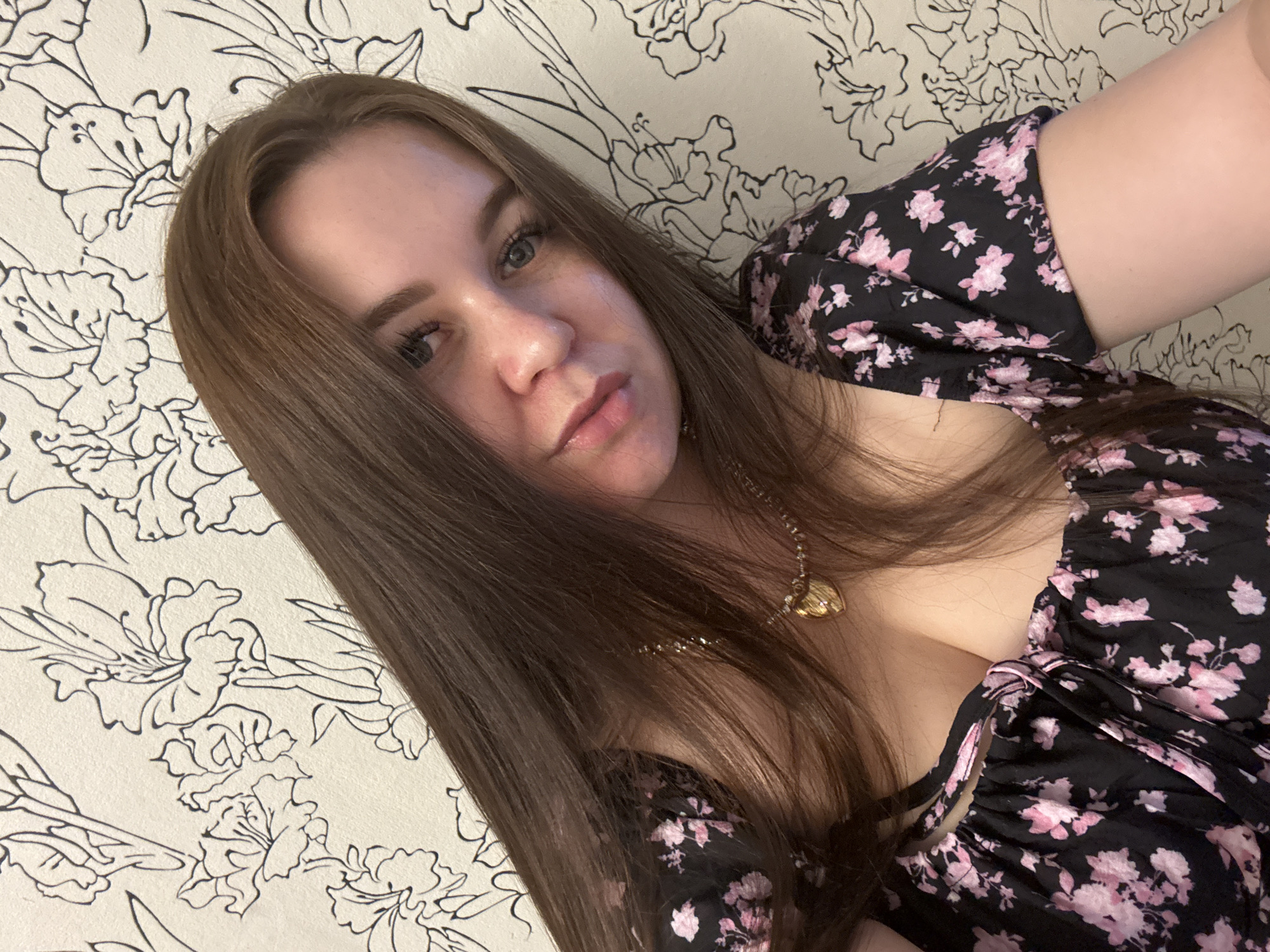 The image shows a close-up, overhead shot of a young woman with long, brown hair. She is wearing a black dress with a pink floral pattern and a gold necklace with a heart-shaped pendant. Her hair is partially obscuring her face, but her blue eyes and lips are visible. She appears to be looking towards the camera with a neutral expression. The background is a wallpaper with a black floral design on a light-colored surface. The lighting suggests it is daytime, and the indoor setting is implied by the wallpaper. The specific location is Chaikovsky, Russia, but no further environmental details are discernible. The image captures a portrait of the woman, with no particular activity or event taking place.