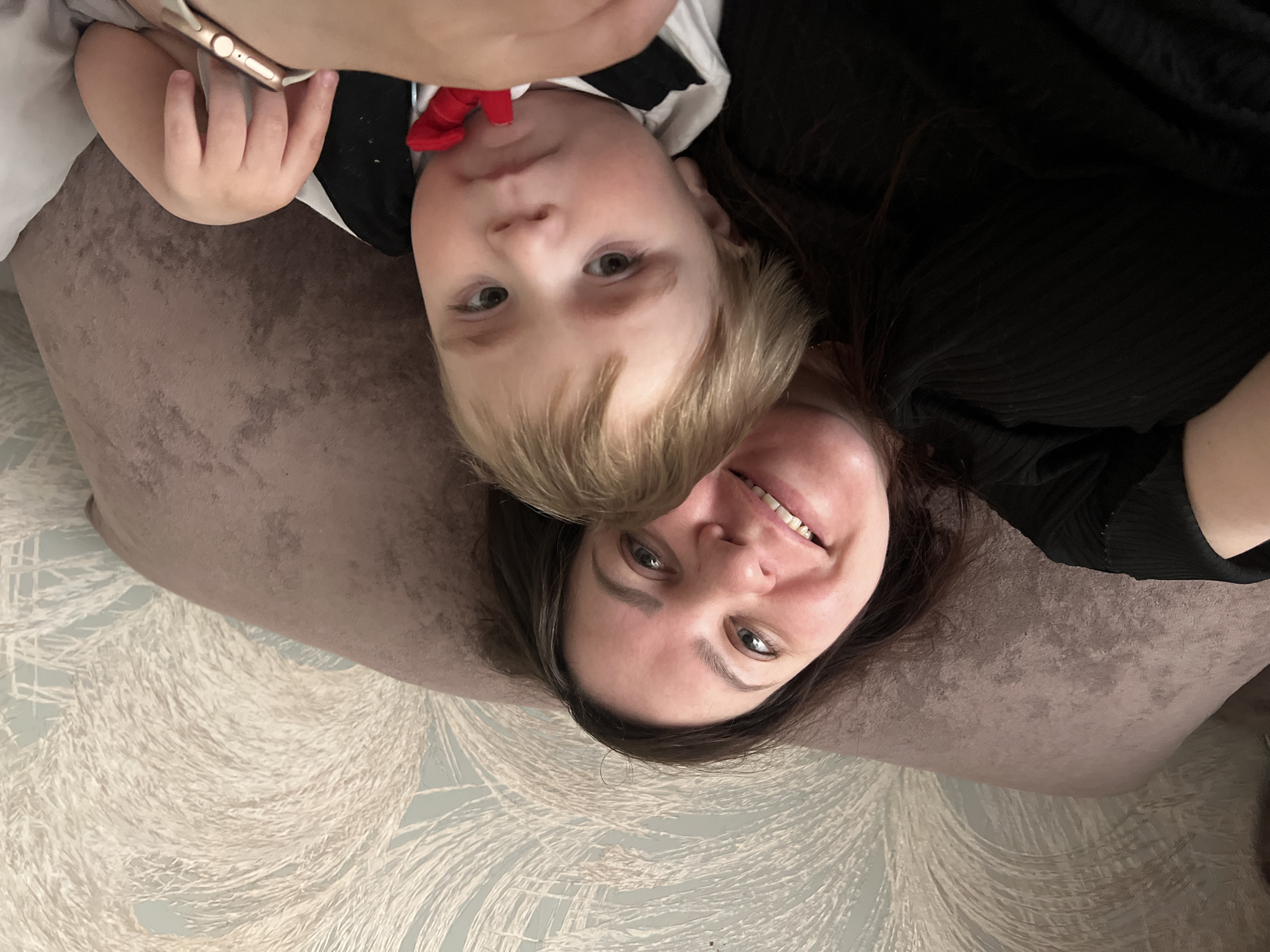 The image shows a selfie of a woman and a young child. The child, wearing a white shirt with a black vest and red bowtie, is positioned above the woman, whose face is near the bottom of the frame. The background consists of a patterned surface, possibly wallpaper or a fabric design, and a portion of a pillow. The picture is taken indoors.
The location in the city of Noviy, Russia cannot be determined with the information provided.