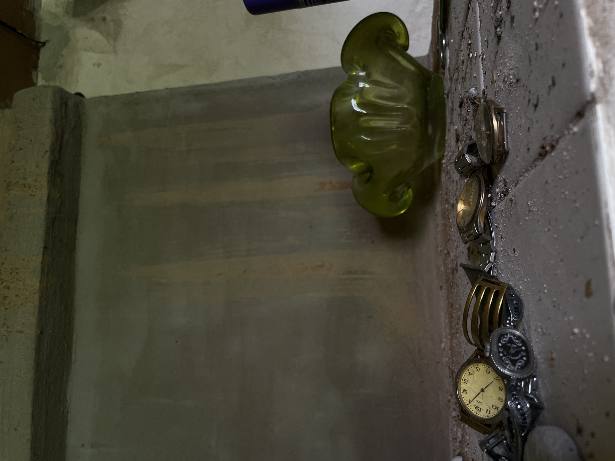 The image depicts an interior scene, likely within a building. The foreground is cluttered with several wristwatches, some appearing vintage, scattered across a dusty, textured surface that resembles a shelf or countertop. The watches vary in design, with visible faces and bands. Among the watches are other small metal objects, possibly jewelry or accessories, also showing signs of age and disuse.
In the midground, to the right of the watches, sits a vibrant green glass ashtray or bowl with a distinctive, ruffled rim. Its shape is organic and reminiscent of a flower. Behind the ashtray, the wall is rendered in a muted, light gray with vertical streaks of yellow and reddish-brown, suggesting possible water damage or staining.
The background consists of a concrete structure, possibly a fireplace or architectural element, with a rough, textured surface. Above this, a dark wooden beam is visible, indicating the ceiling or upper structure of the room. The overall lighting is dim, casting subtle shadows and highlighting the dusty, neglected state of the objects and the environment. The scene evokes a sense of abandonment or disarray.