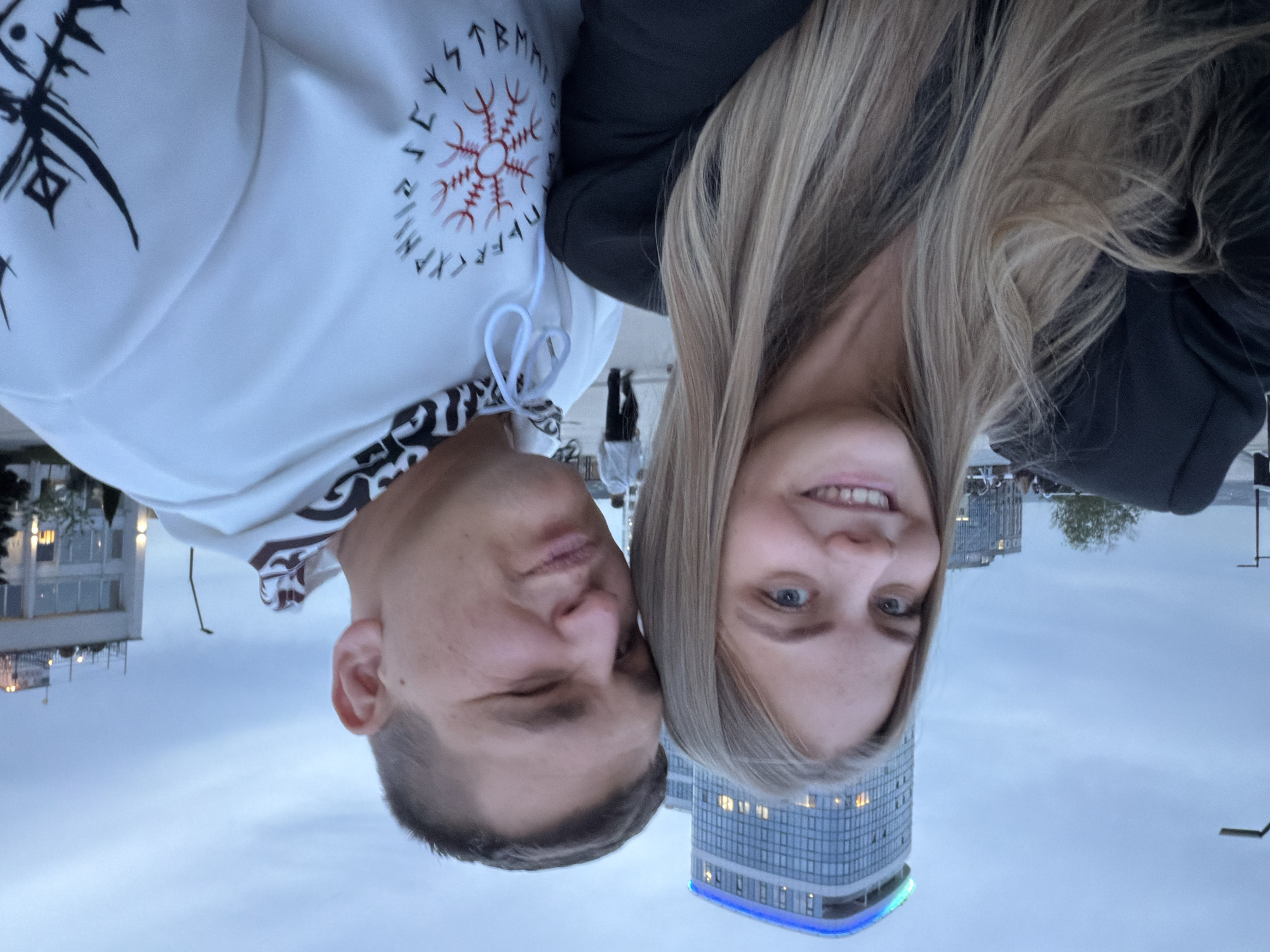 A man and woman pose closely together in an outdoor selfie. The background features buildings and a cloudy sky. The woman has long, blonde hair and is smiling, while the man has short, dark hair. He is wearing a white hoodie with black and red Viking-inspired designs, including runes and a Vegvísir symbol. The image appears to be upside down. The building in the background with the blue light is the “Torch” building in the city of Izhevsk, Russia.