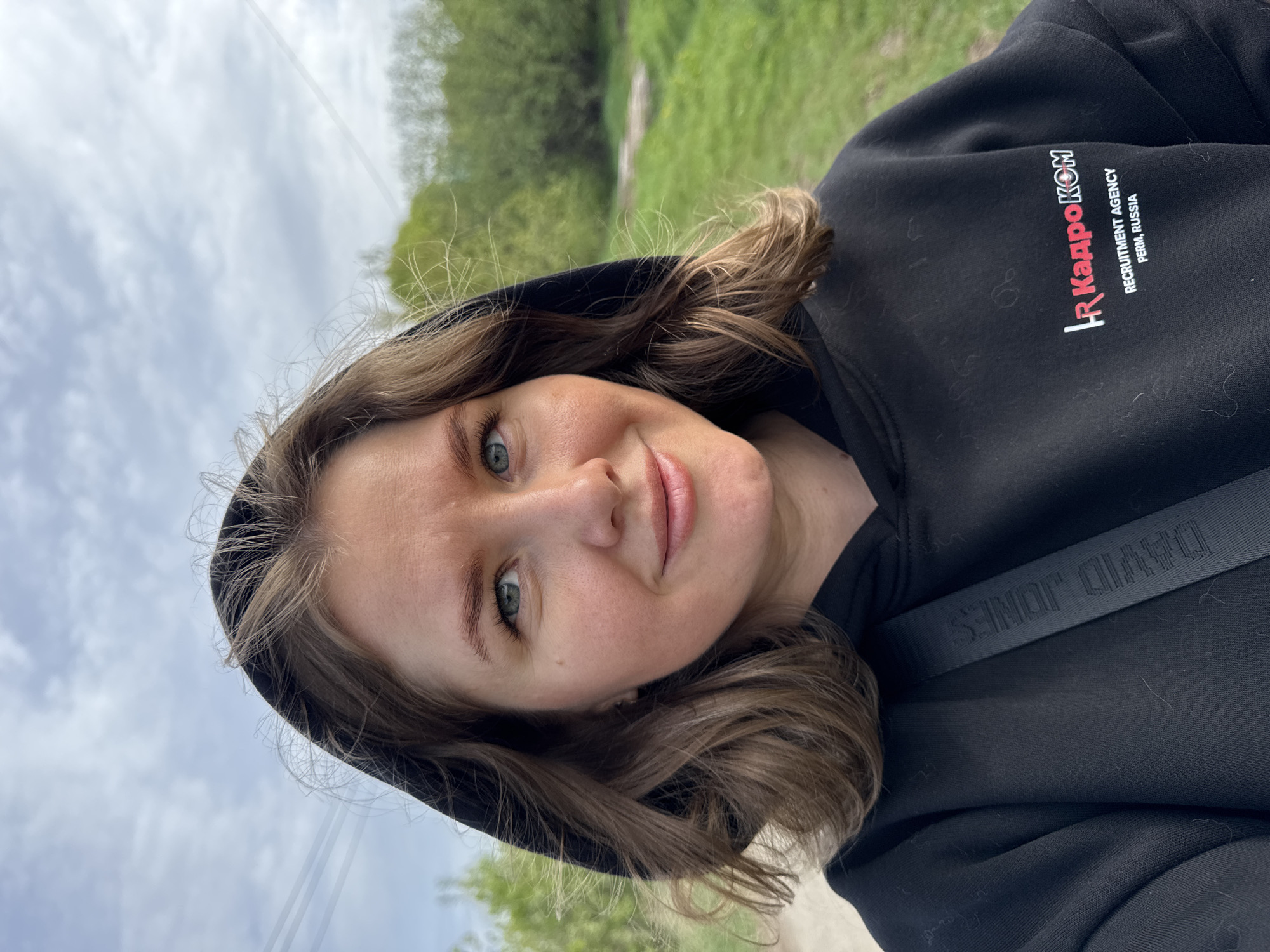 A woman is shown in a close-up selfie outdoors. She has fair skin, light brown hair with waves, and light-colored eyes. She is wearing a black hooded sweatshirt with the text "HR Кадроком Recruitment Agency Perm, Russia" printed on the chest in red and white lettering. The hood is up, framing her face. A black strap with "DAVID JONES" printed on it is visible across her torso. The background is blurry and appears to be a green grassy area with trees. Overhead wires are faintly visible against a cloudy sky. The photo is slightly rotated counter-clockwise. The scene is in Горный, Russia.